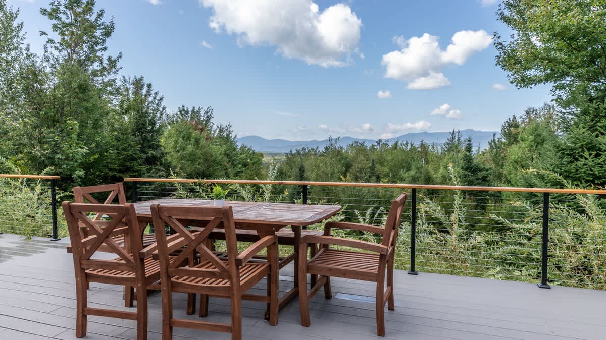 Outdoor deck furnished with a wooden dining table and four chairs, providing a scenic overlook of a vast green forest and distant mountains under a bright sky.