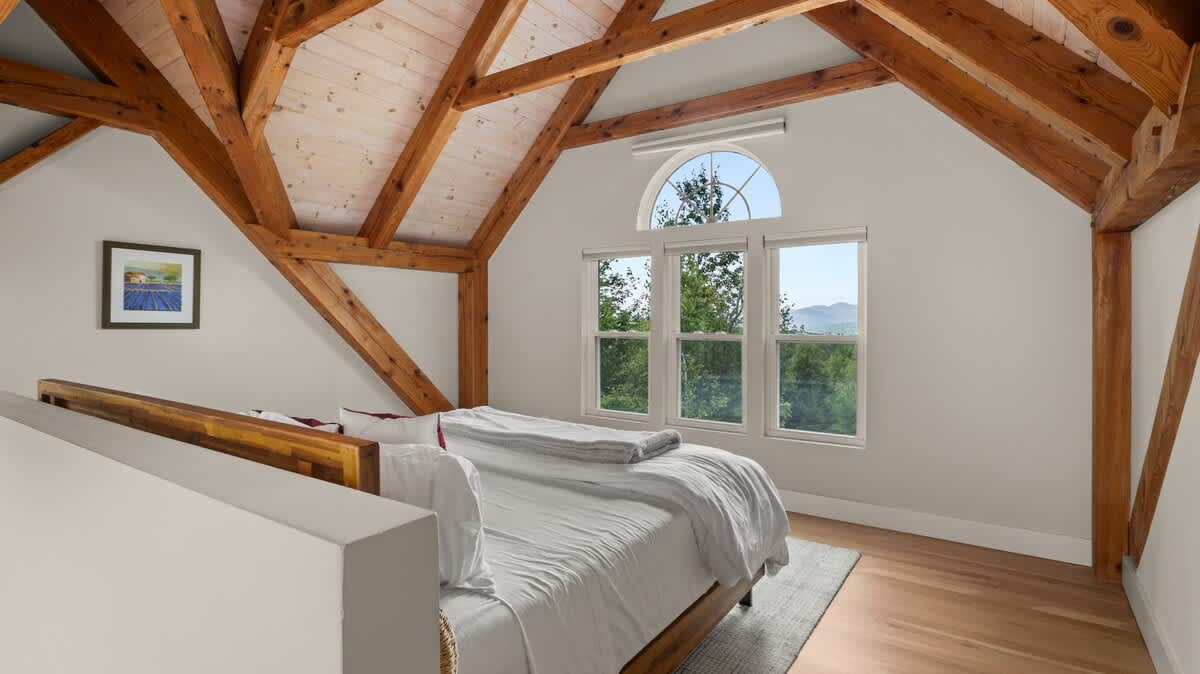 A bright bedroom features a bed with crisp white bedding, set against a light gray wall. Exposed wooden beams create a vaulted ceiling, and a large window with an arched top offers scenic views of lush trees and distant mountains.