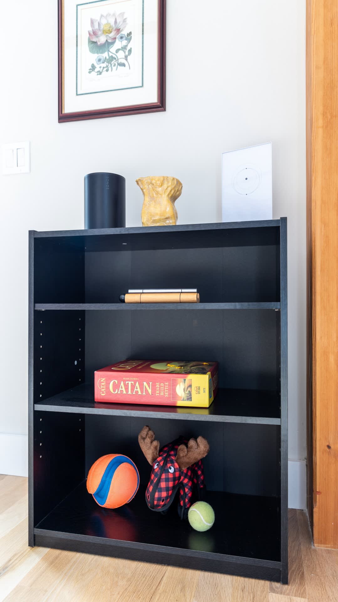 Black three-shelf bookcase filled with board games, a plush moose toy, and a colorful ball, suggesting a family-friendly space. A framed floral print hangs above the shelf on a light gray wall.
