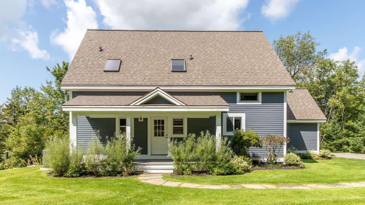 Front exterior view of a charming two-story house featuring dark blue siding, a light brown shingle roof with skylights, and a welcoming covered porch with white trim. The property is set on a manicured green lawn with lush landscaping.
