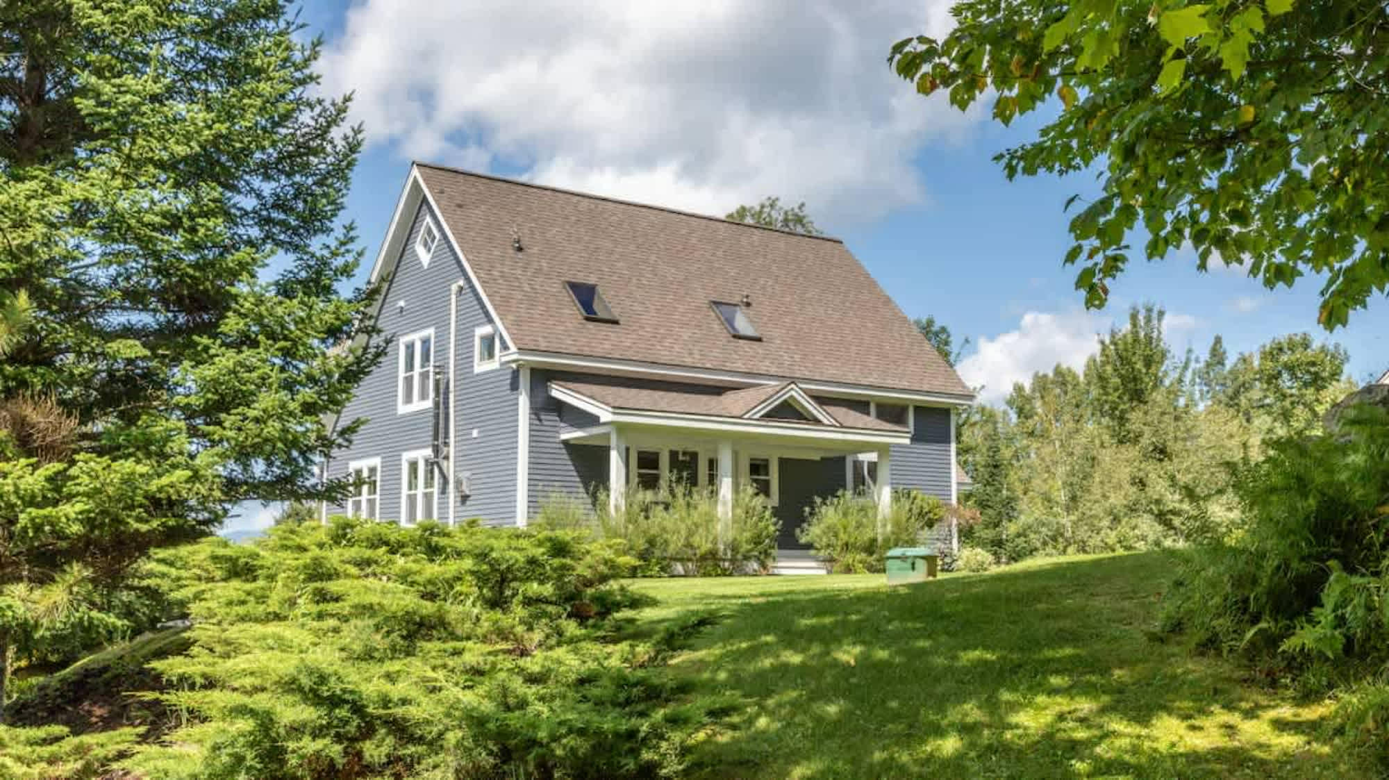 Front view of the blue house with white trim, a covered porch, and multiple windows, surrounded by manicured green bushes and tall trees under a bright sky.