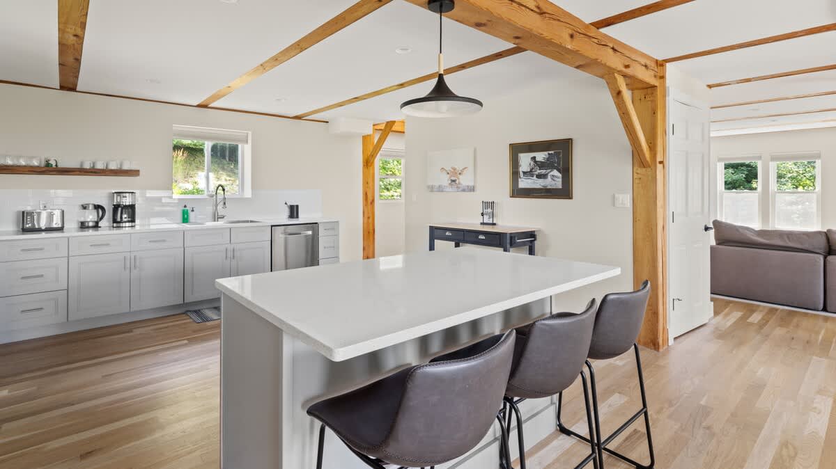 Open-concept kitchen and dining area featuring a large white island with three bar stools, light gray cabinets, and exposed wooden beams, leading into a living area with a sofa.