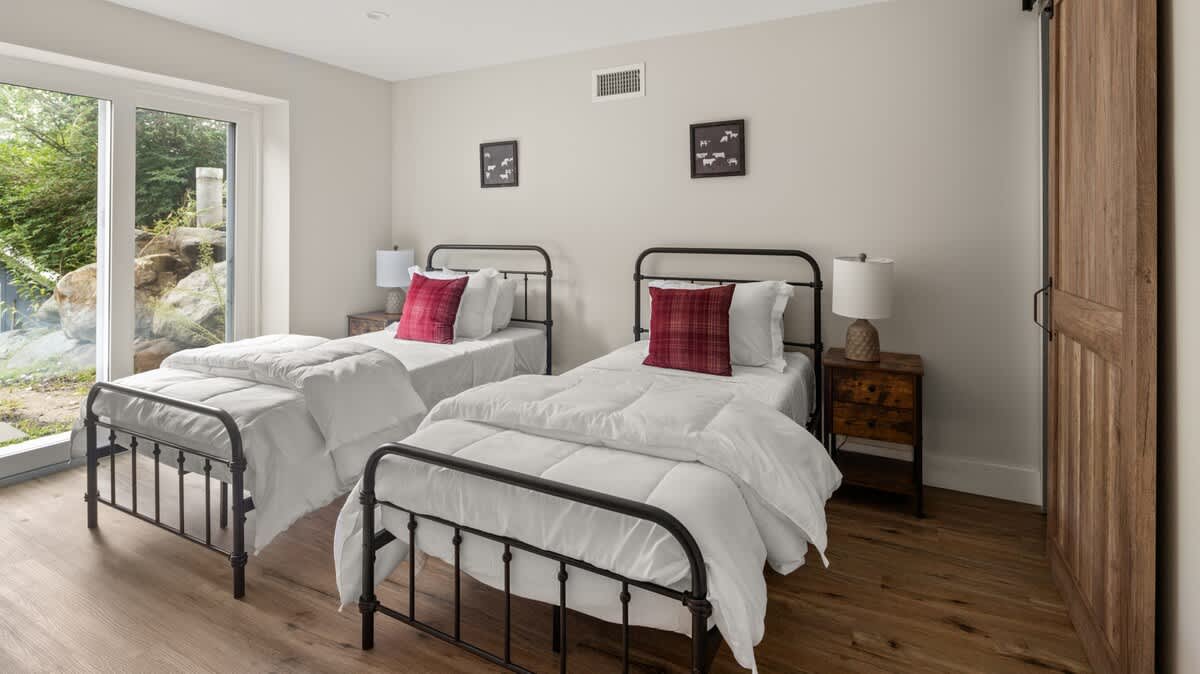 Bright bedroom featuring two black metal-frame twin beds with crisp white bedding and red plaid accent pillows, set on light wood flooring. A large glass door opens to an outdoor area with natural rock and greenery.