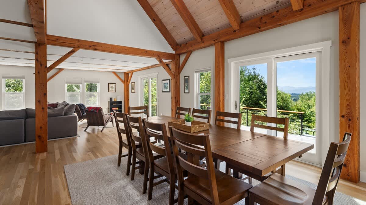 Bright dining area with a large wooden table seating ten, surrounded by wooden chairs, set on a light area rug. Large sliding glass doors offer views of lush greenery and distant mountains.