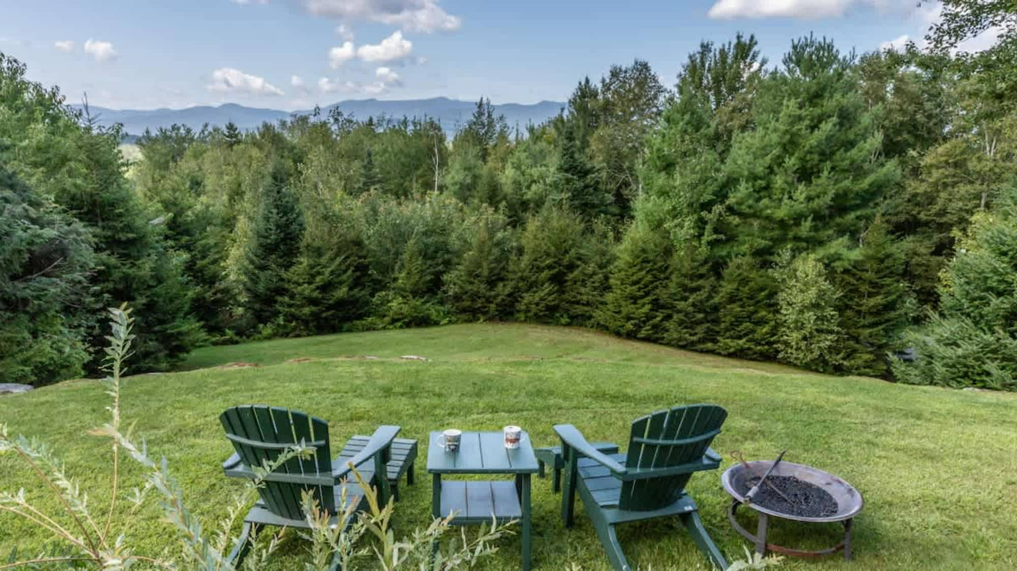 Grassy backyard area with two green Adirondack chairs, a small table, and a metal fire pit, set against a backdrop of dense green trees and distant mountains under a cloudy sky.