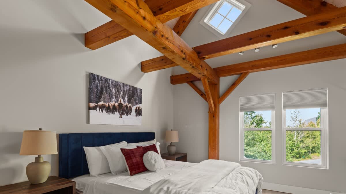 A wide view of the bedroom highlights a queen bed with a blue velvet headboard, flanked by two nightstands with lamps. A large artwork of bison hangs above the bed, while exposed wooden beams crisscross the ceiling. Two windows offer views of trees and mountains, complemented by a skylight.