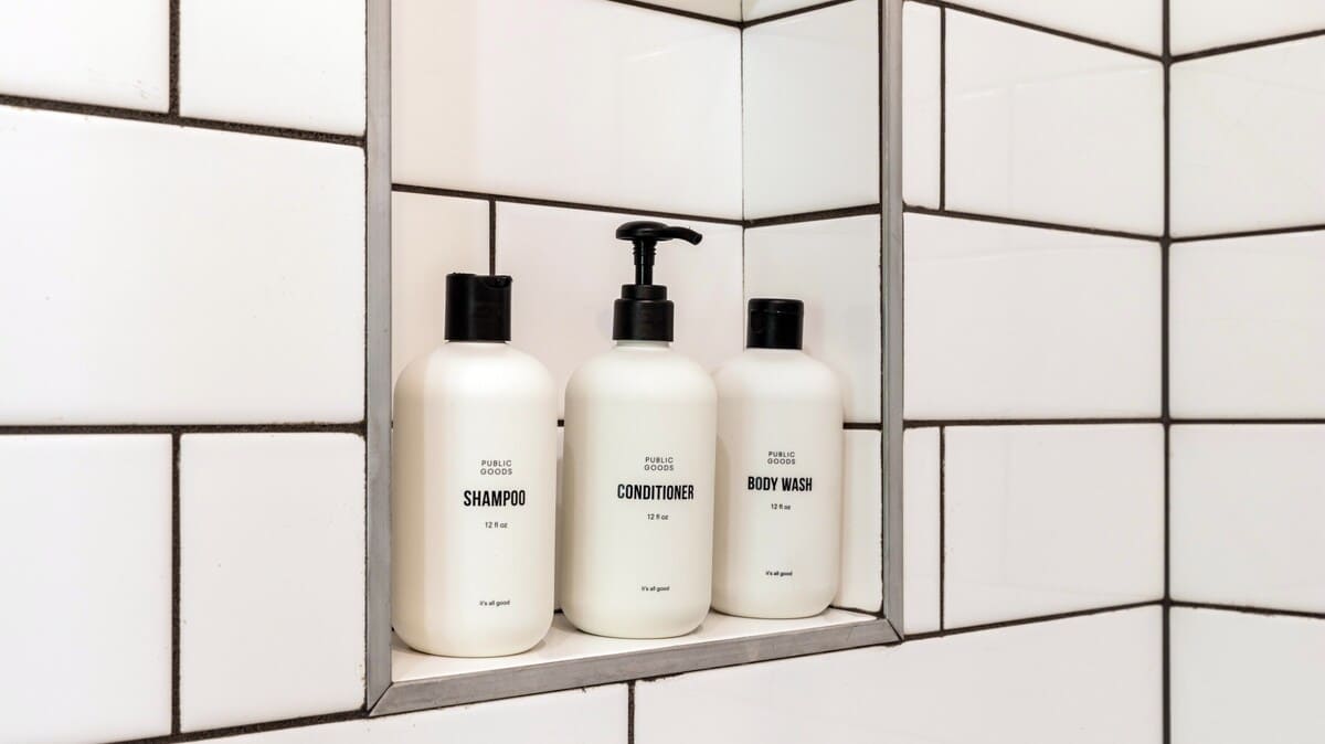 A close-up view inside a shower niche reveals three white bottles of 'Public Goods' brand shampoo, conditioner, and body wash, neatly arranged against a backdrop of white subway tiles with dark grout.