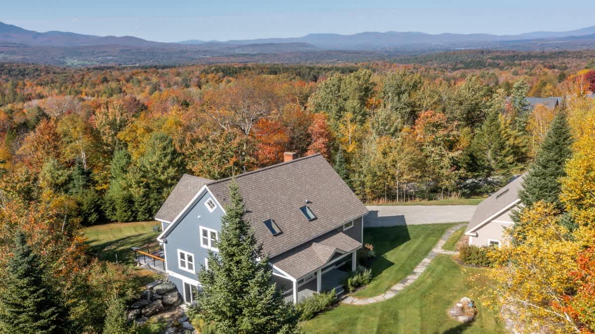Closer aerial view of the main blue-sided house with a brown roof, surrounded by a mix of evergreen and vibrant autumn-colored trees. A second smaller building and a winding pathway are visible, with distant mountains under a clear sky.