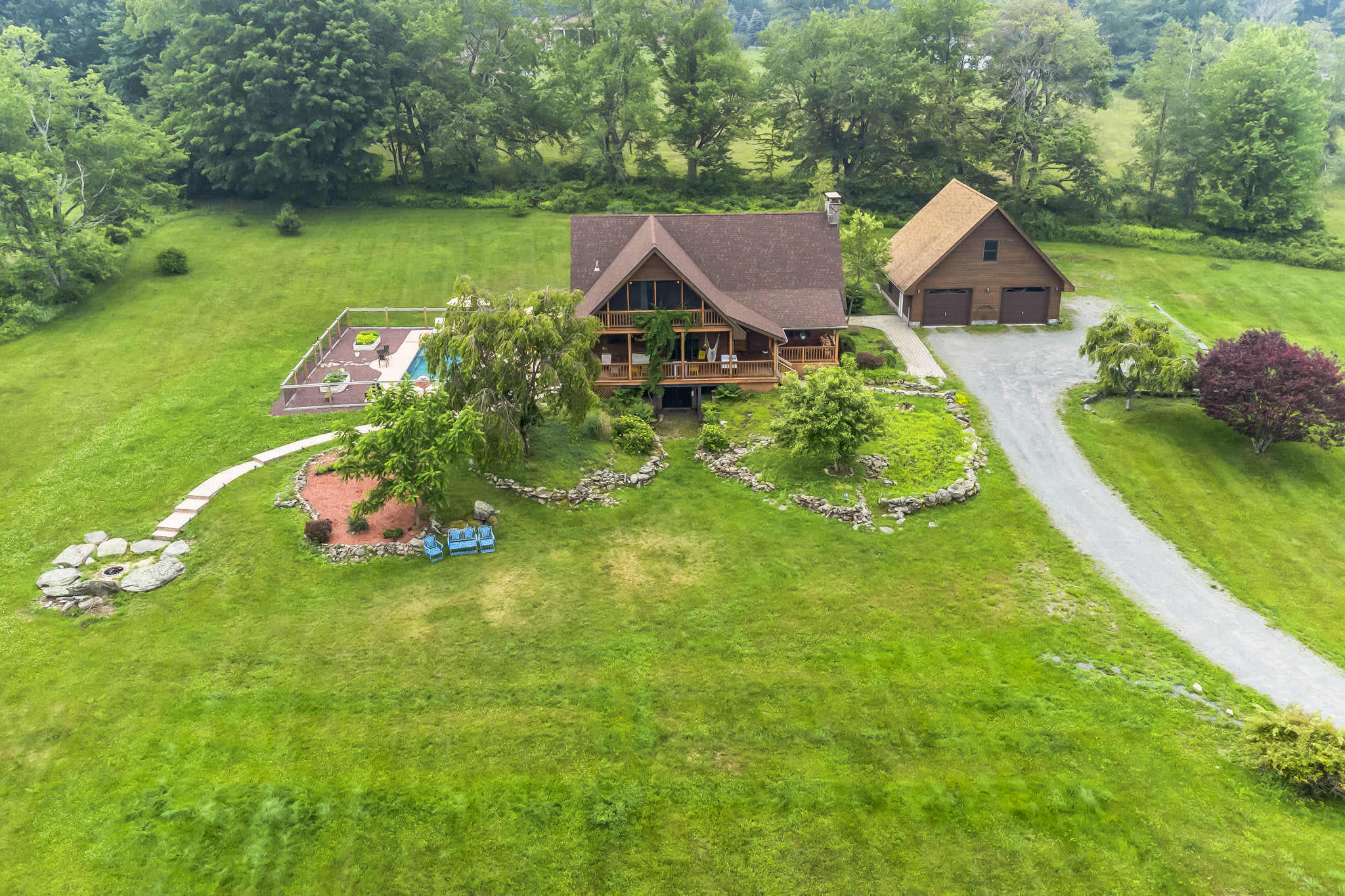 An aerial view showcases a large A-frame style house with a brown roof and wooden siding, surrounded by vast green lawns and mature trees. A fenced swimming pool area and a separate garage structure are also visible on the expansive property.
