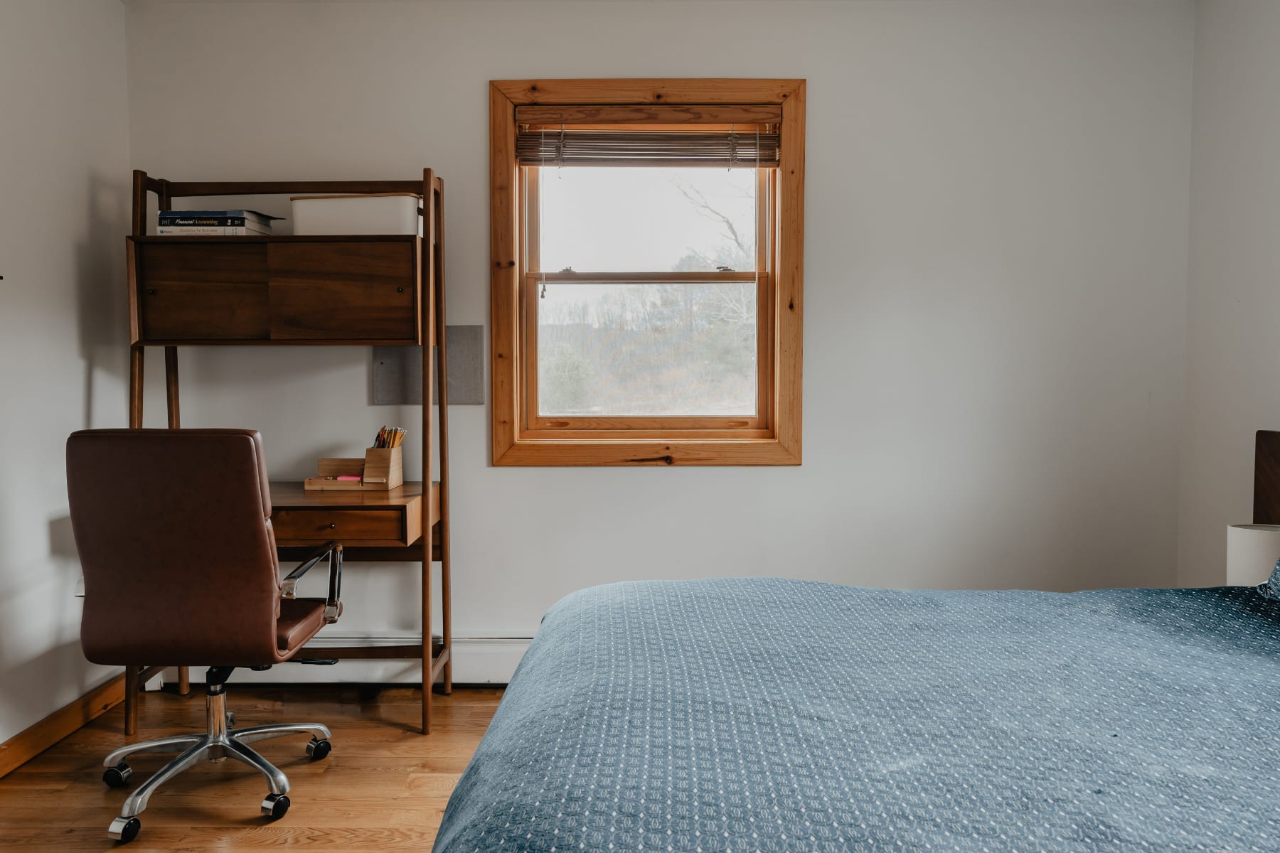 A bright bedroom with light gray walls and wooden floors, featuring a wooden desk and shelving unit with a brown office chair, positioned next to a large window with a wooden frame and woven blinds.
