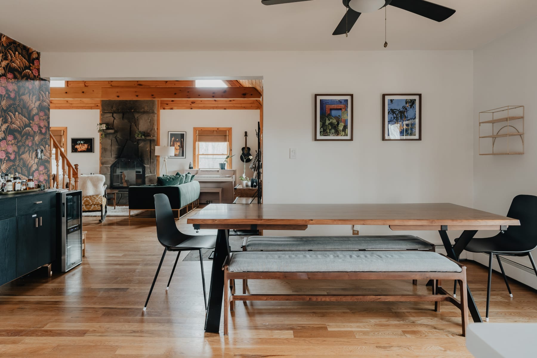 A wide view of the dining area with a long wooden table and benches, offering a glimpse into the adjacent living room with its fireplace and sofas. The open layout connects the spaces, featuring hardwood floors and a modern aesthetic.