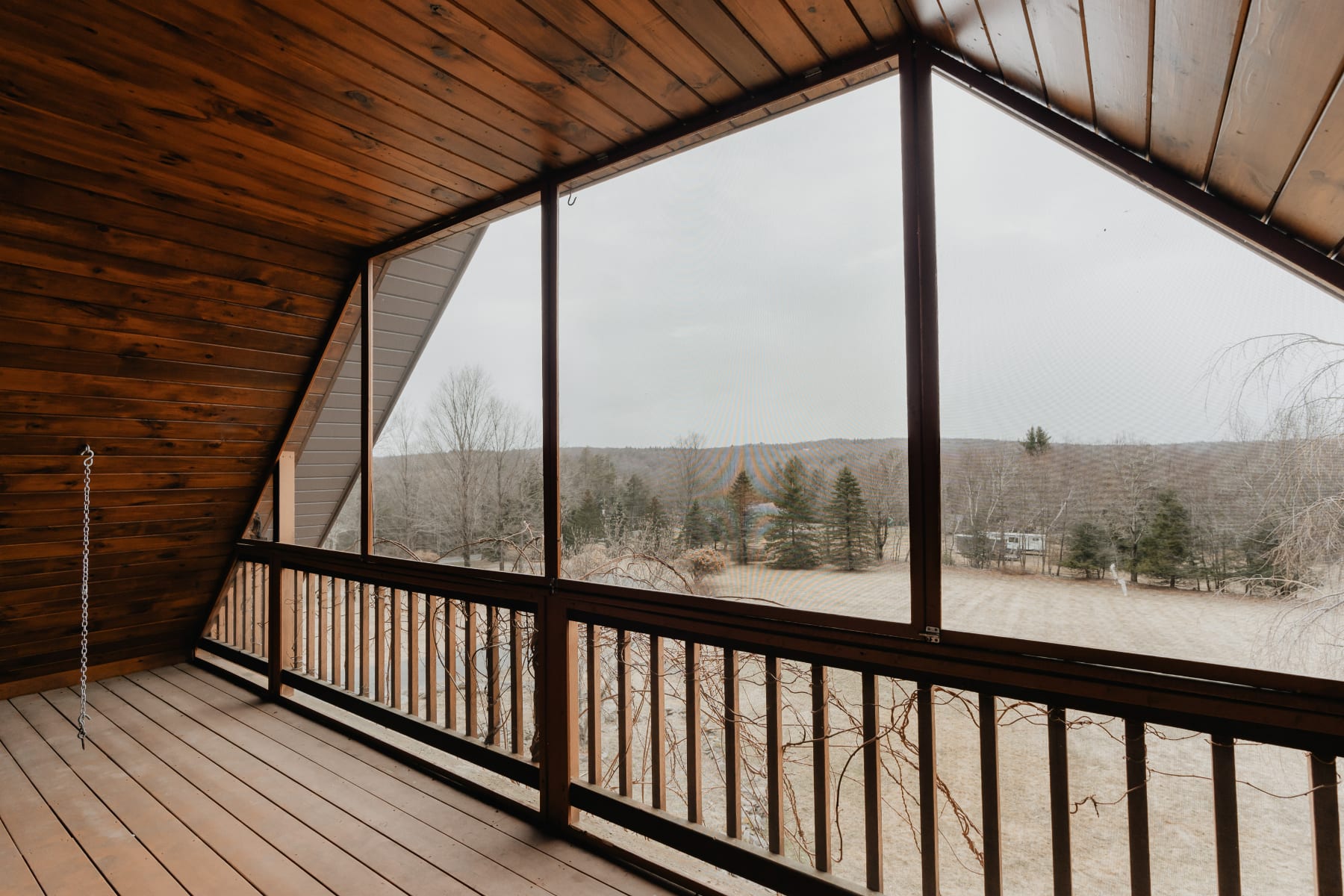 A screened-in balcony with a sloped wooden ceiling and floor, offering a view of a wooded landscape with bare trees and a distant house. A wooden railing separates the interior from the screened exterior.