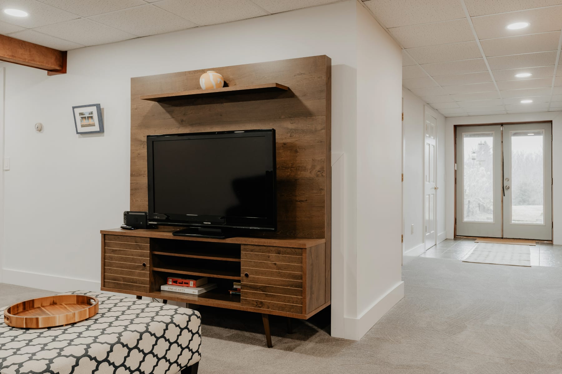 A modern living room with a large flat-screen TV mounted on a wooden media console with a floating shelf. A patterned ottoman is in the foreground, and the room has light gray carpet and white walls.