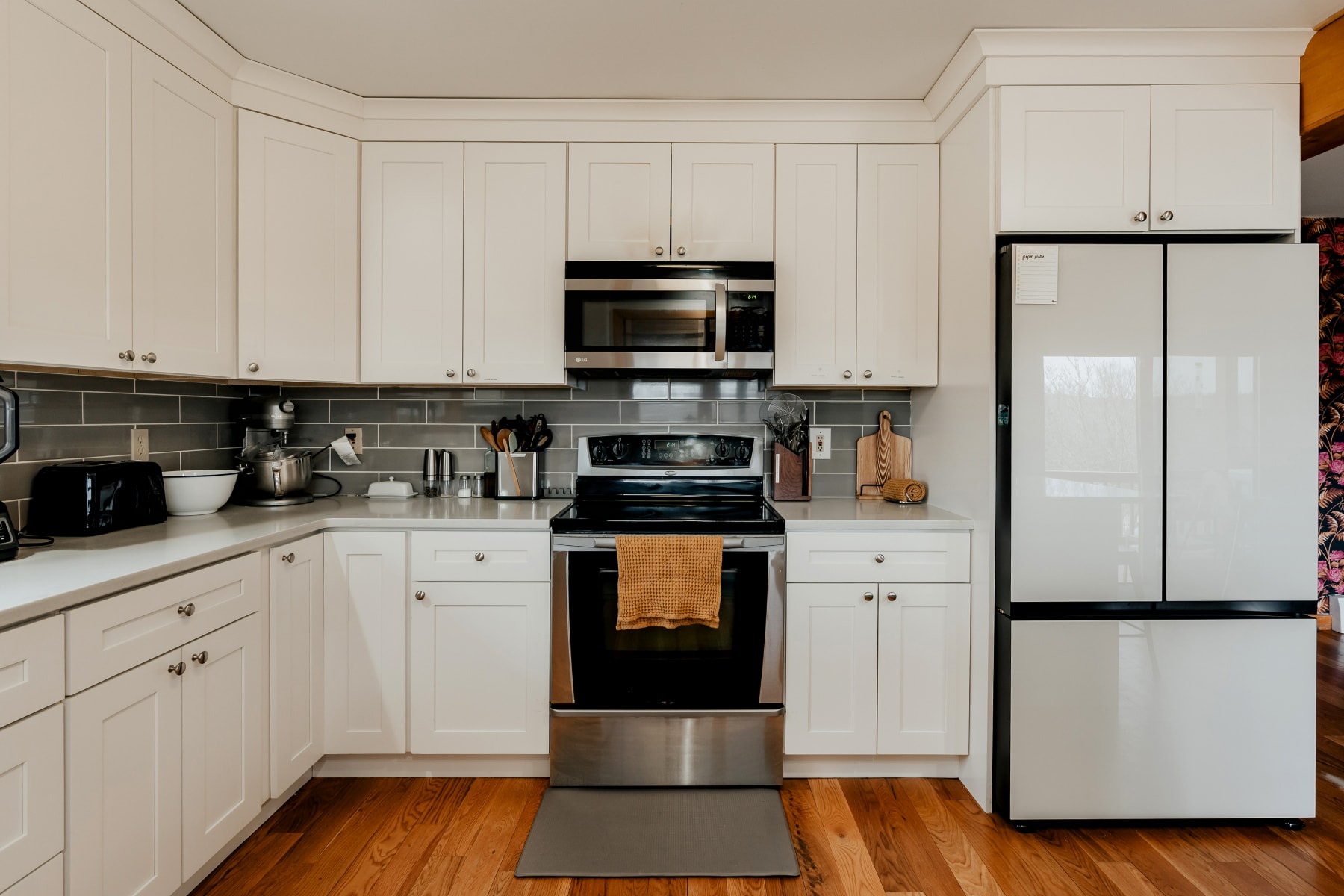 A well-equipped kitchen featuring white shaker cabinets, light gray subway tile backsplash, and light gray countertops, with a stainless steel oven/range, microwave, and a large black and white refrigerator.