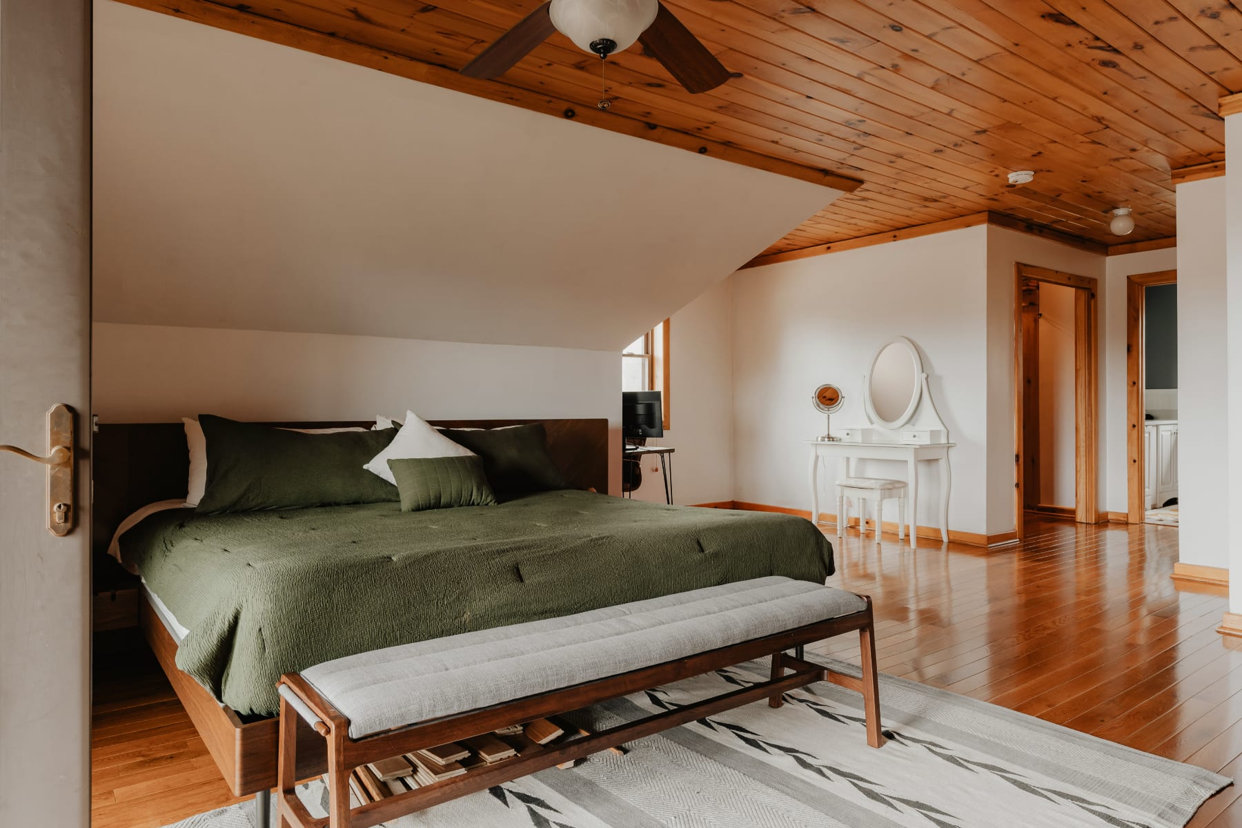 A cozy attic bedroom with a queen-sized bed featuring dark green bedding, a wooden headboard, and a light gray bench at the foot. The room has warm wooden floors and a sloped wooden ceiling, with a white vanity and mirror in the background.