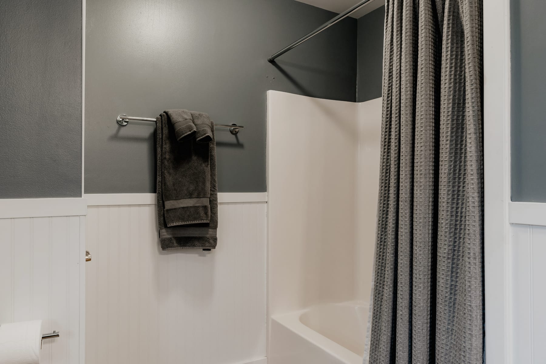 A section of a bathroom with dark gray walls and white wainscoting, featuring a white shower/tub combo with a gray waffle-weave shower curtain. A towel rack with gray towels is mounted on the wall.
