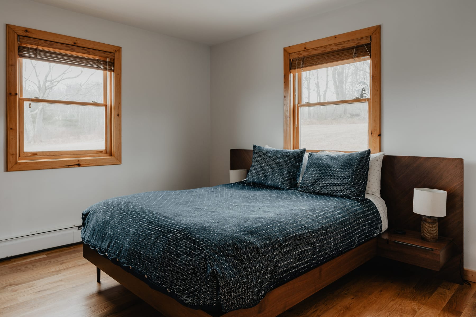 A cozy bedroom with light gray walls and warm wooden floors, featuring a queen-sized bed with a dark blue patterned quilt, two large windows with wooden frames, and a modern wooden headboard with integrated nightstands.