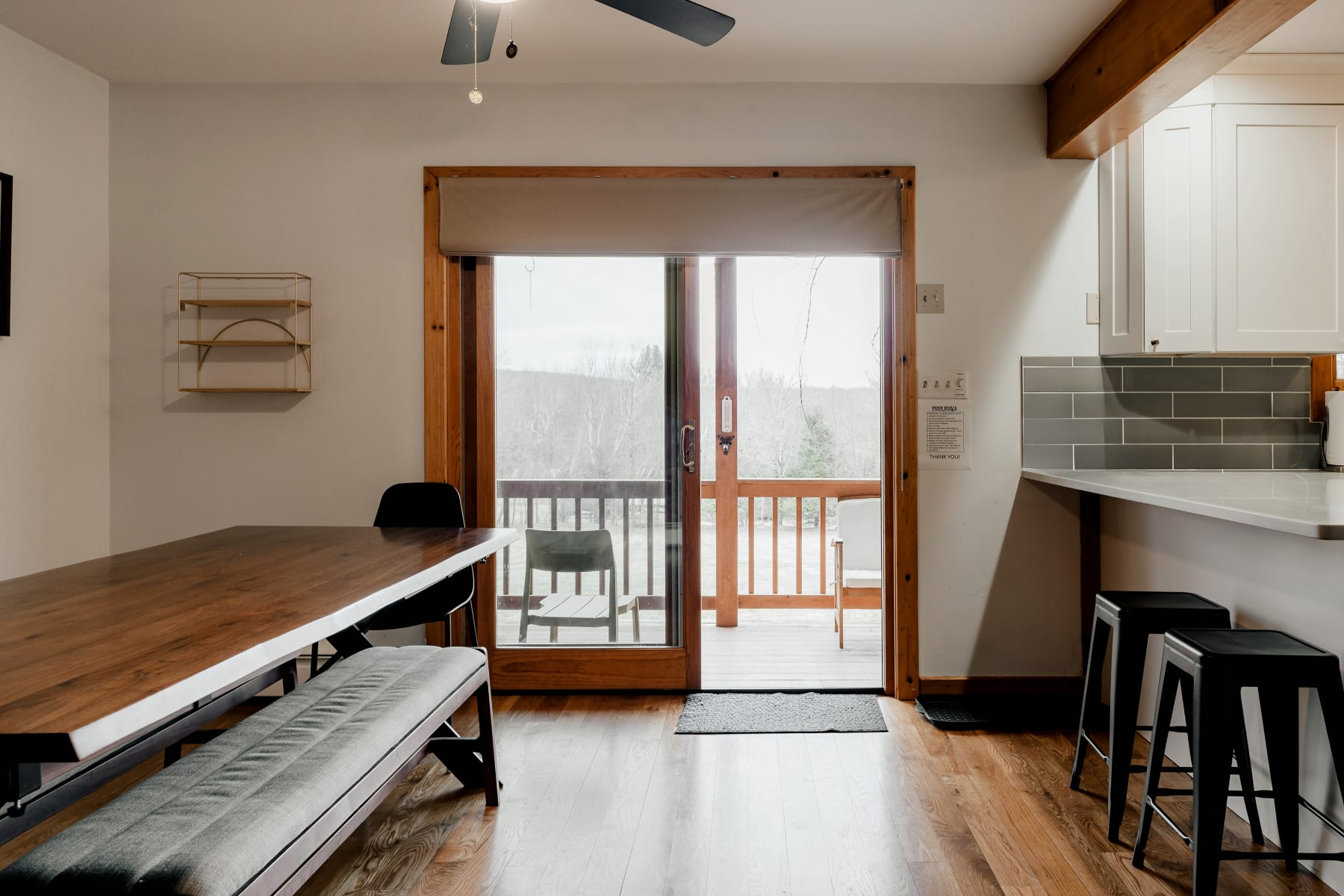 The dining area with a wooden table and benches, featuring a large sliding glass door that opens to an outdoor deck, offering views of the surrounding landscape. The modern kitchen with a breakfast bar is visible to the right.