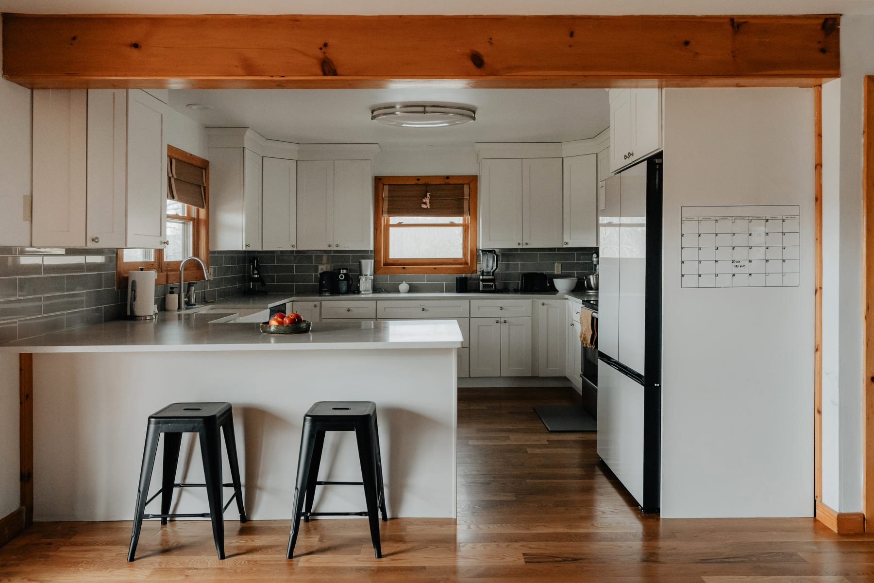 A modern kitchen with white shaker cabinets, light gray subway tile backsplash, light gray countertops, and dark wood floors, featuring a central island with two black bar stools and a stainless steel refrigerator.