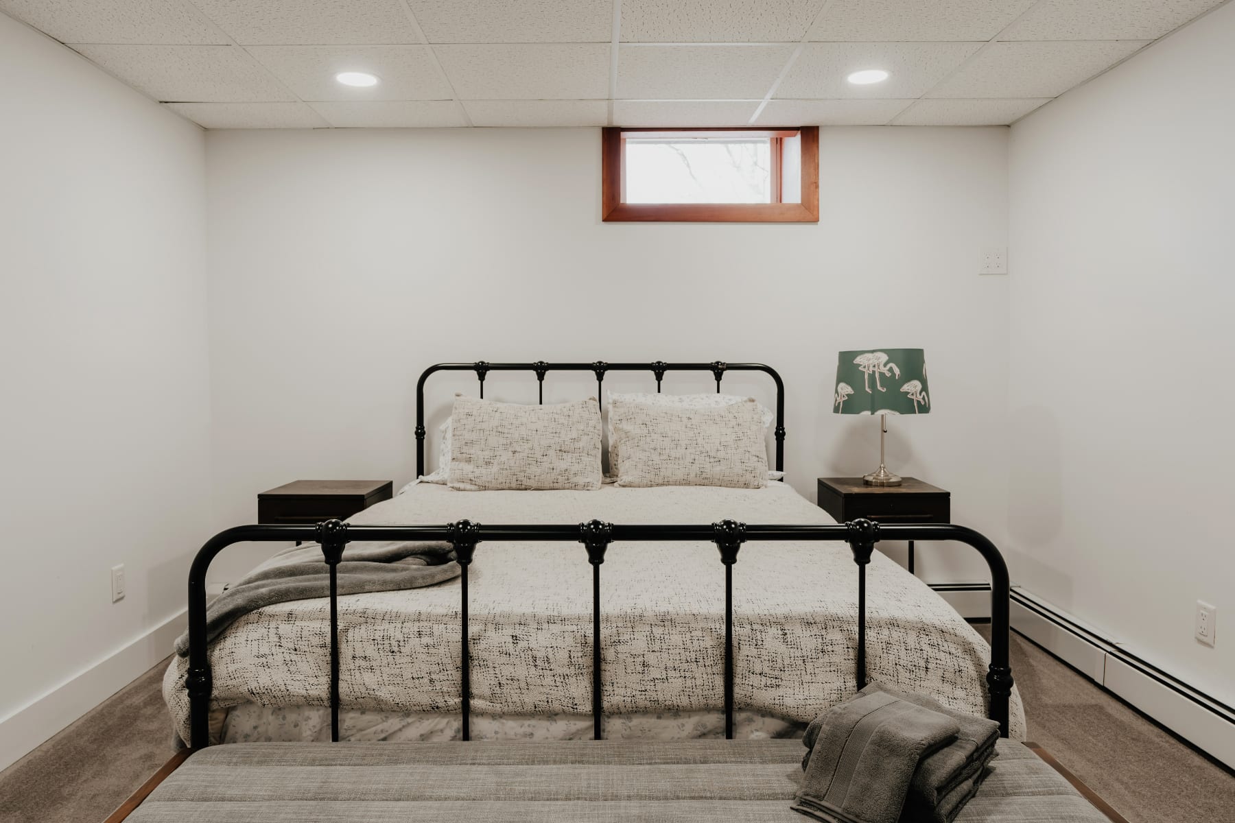 A basement bedroom with a black metal frame queen bed, white and gray patterned bedding, and two dark wood nightstands with lamps. A small window with a wooden frame is high on the white wall, and the room has a drop ceiling with recessed lighting.