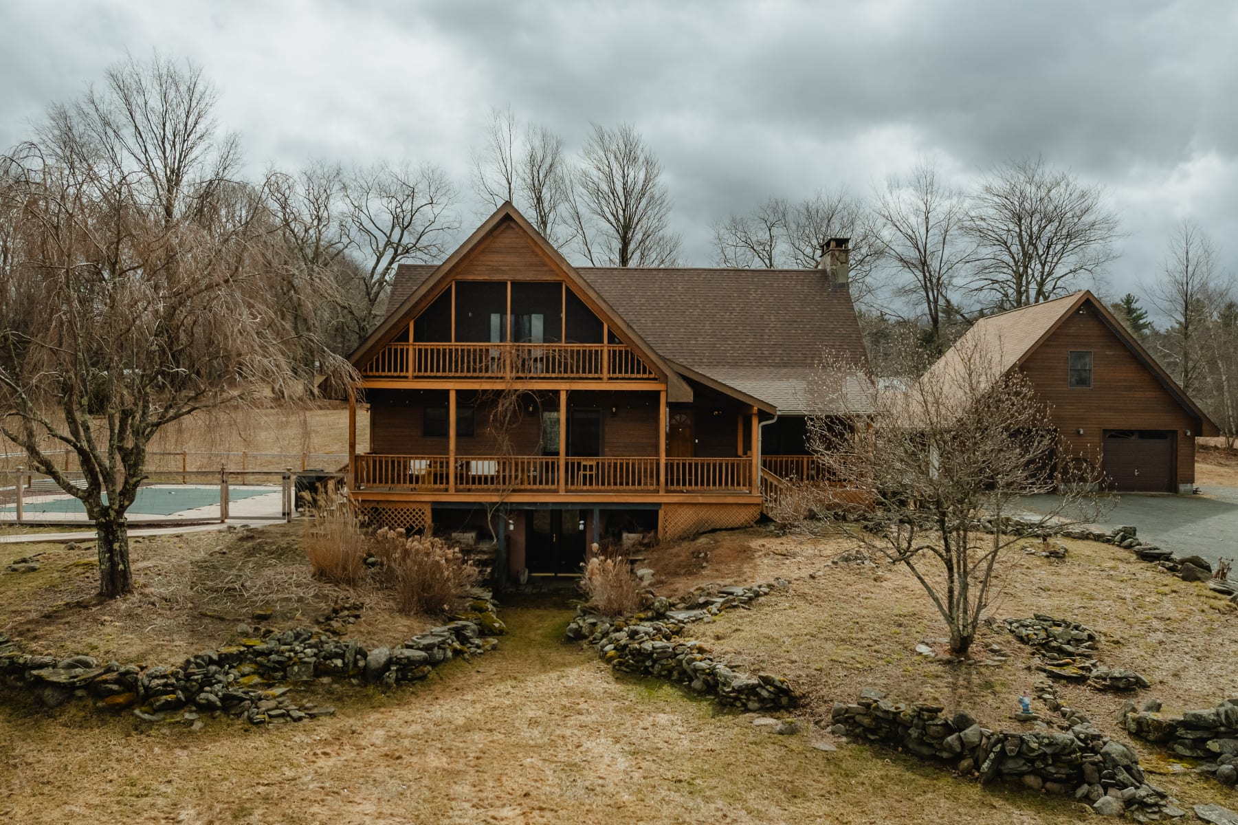 Back exterior of a multi-story wooden house with a screened-in porch and multiple balconies, overlooking a sloped yard with stone walls and bare trees under a cloudy sky.
