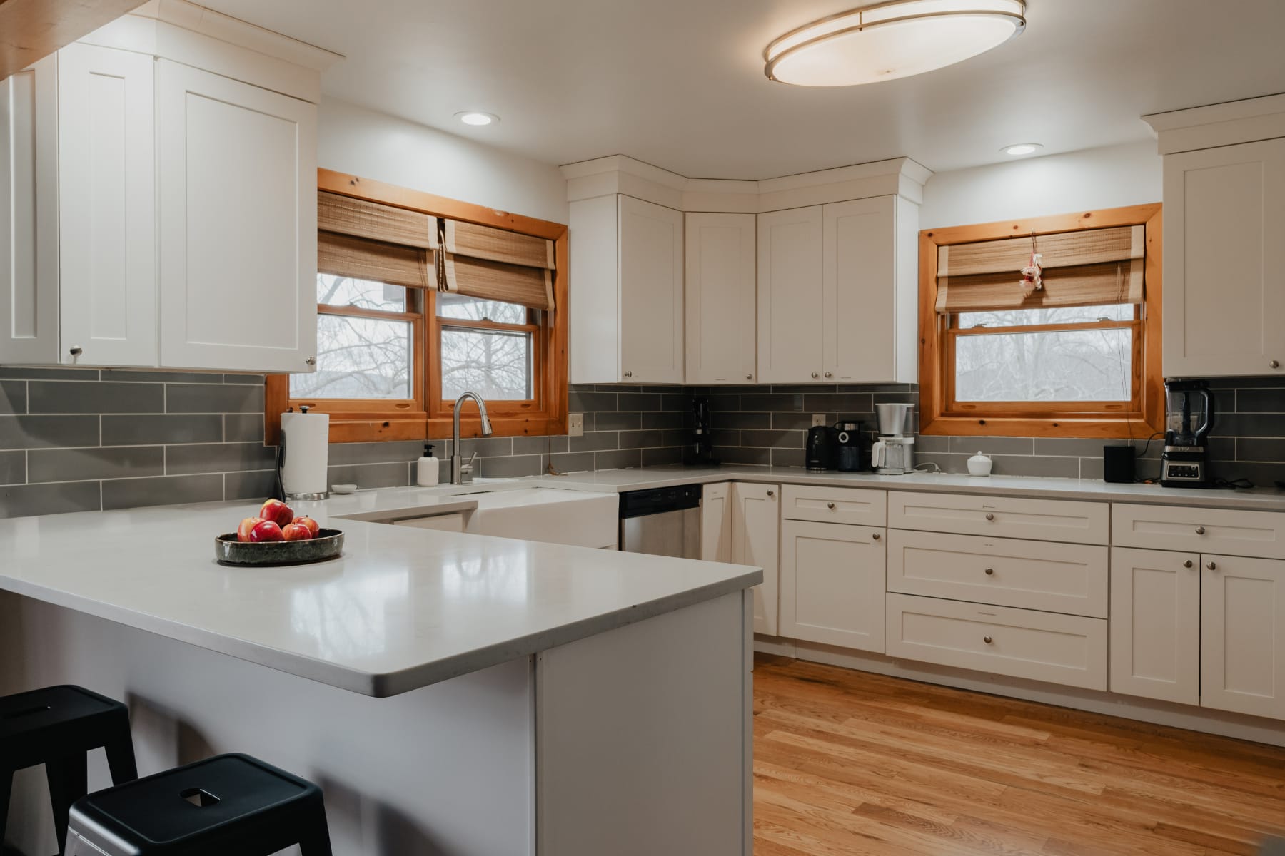 A bright kitchen with white shaker cabinets, light gray subway tile backsplash, and light gray countertops, showcasing a farmhouse sink, stainless steel dishwasher, and two large windows with wooden frames and woven blinds.