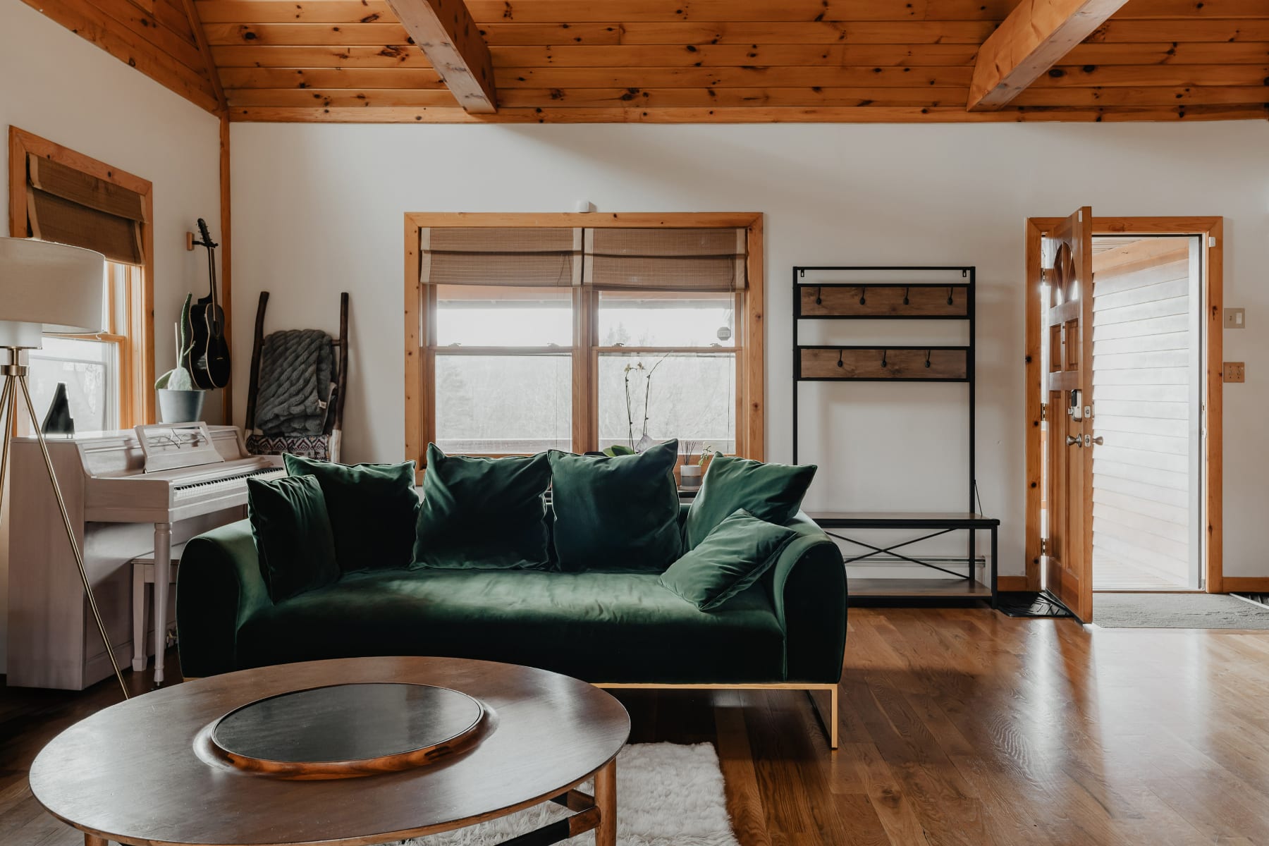 A cozy living room featuring a dark green velvet sofa with gold legs, a round wooden coffee table, and a white upright piano. The room has warm wooden floors, exposed ceiling beams, and a large window with natural light.