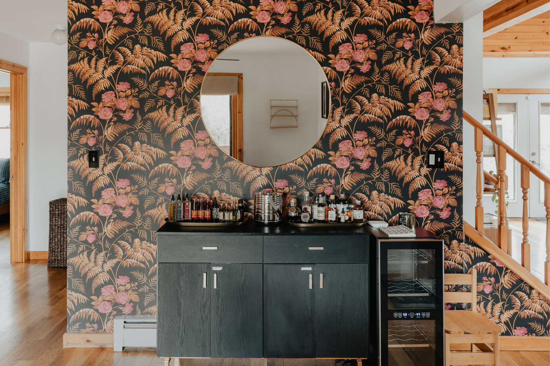 Stylish bar area with dark wood cabinetry, a round mirror, and floral wallpaper, located near a staircase with wooden railings, offering a dedicated space for drinks.