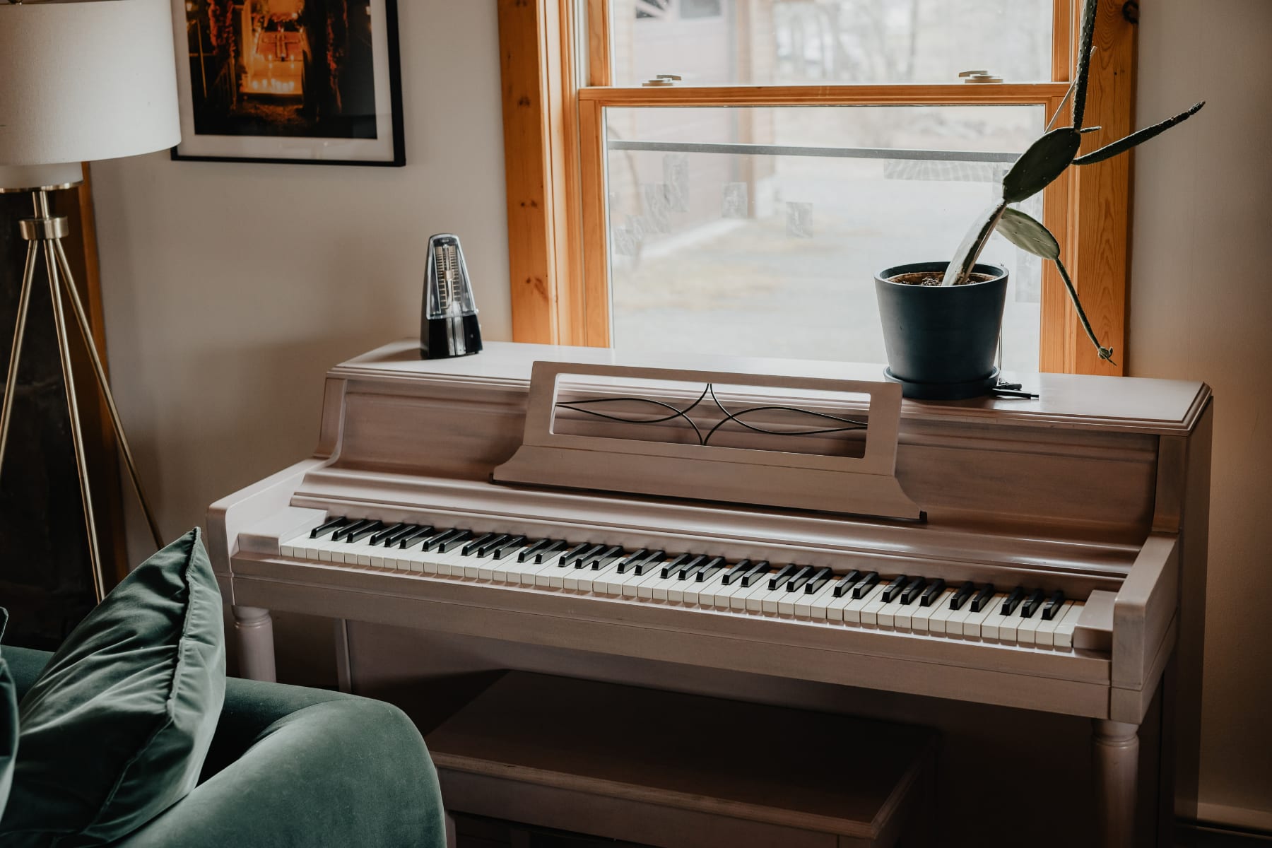 A detailed view of a light-colored upright piano with a metronome on top, positioned by a window. A potted cactus sits on the windowsill, adding a touch of greenery to the musical corner.