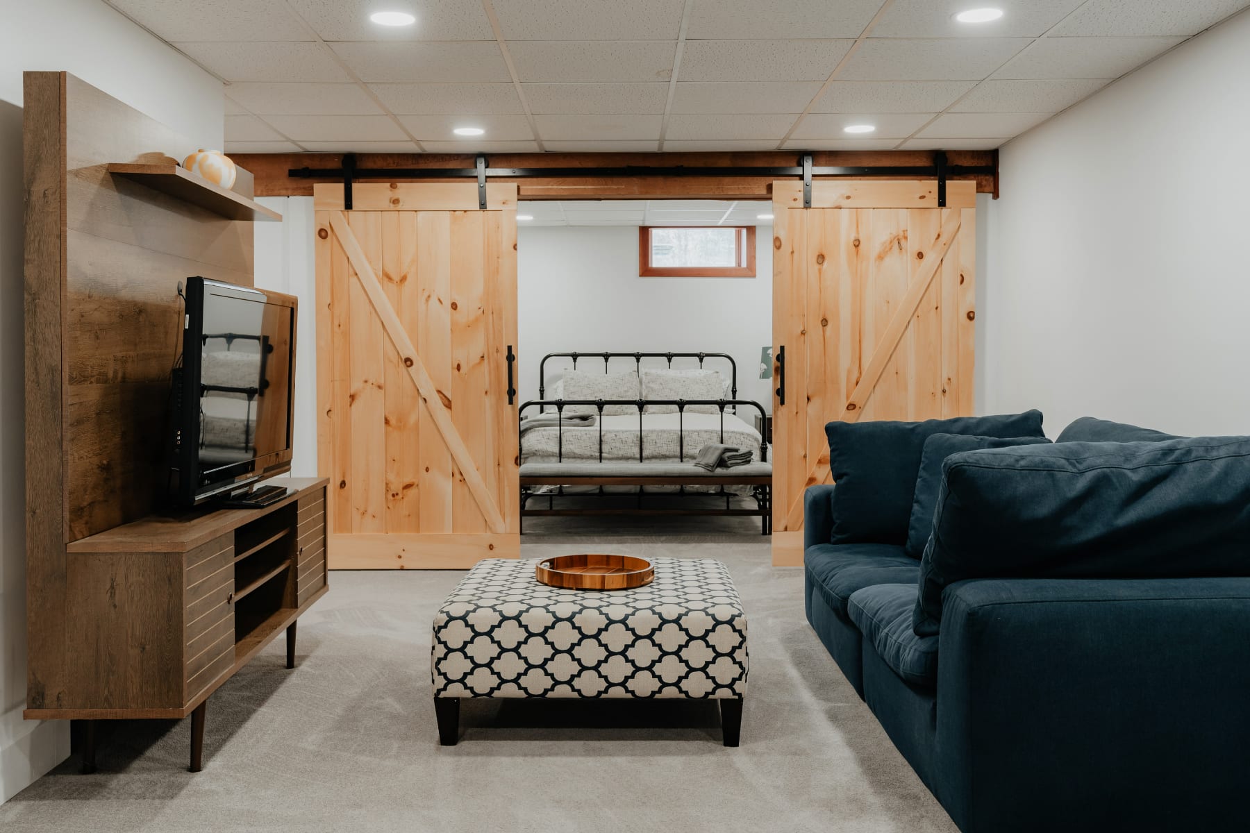 A spacious living area with a navy blue sofa and a patterned ottoman, separated from a bedroom by two rustic wooden barn doors. The room features a light gray carpet and a drop ceiling with recessed lighting.