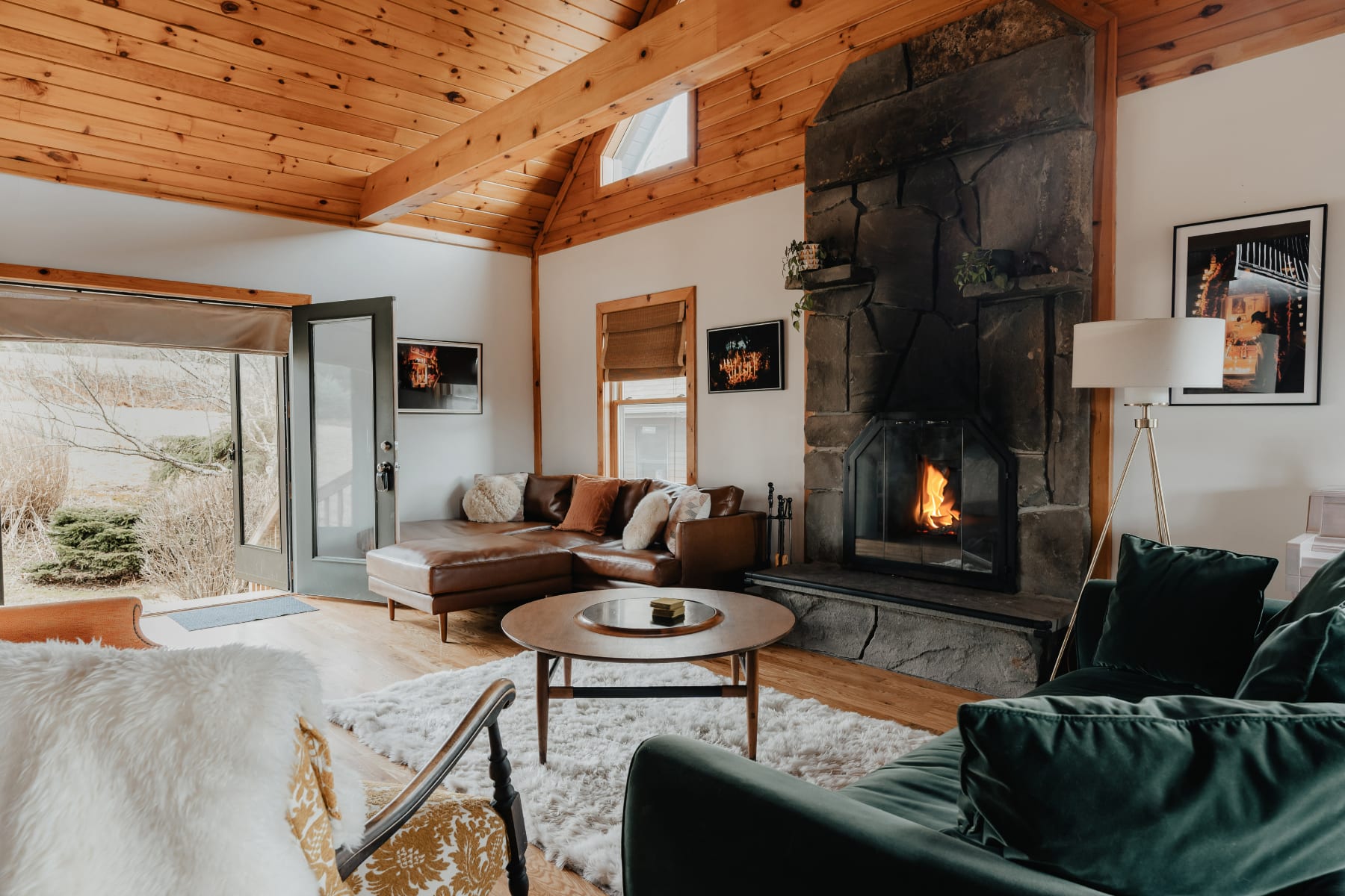 Cozy living room with a large stone fireplace, vaulted wood-paneled ceiling, a brown leather sectional, and a dark green velvet sofa, centered around a round wooden coffee table.