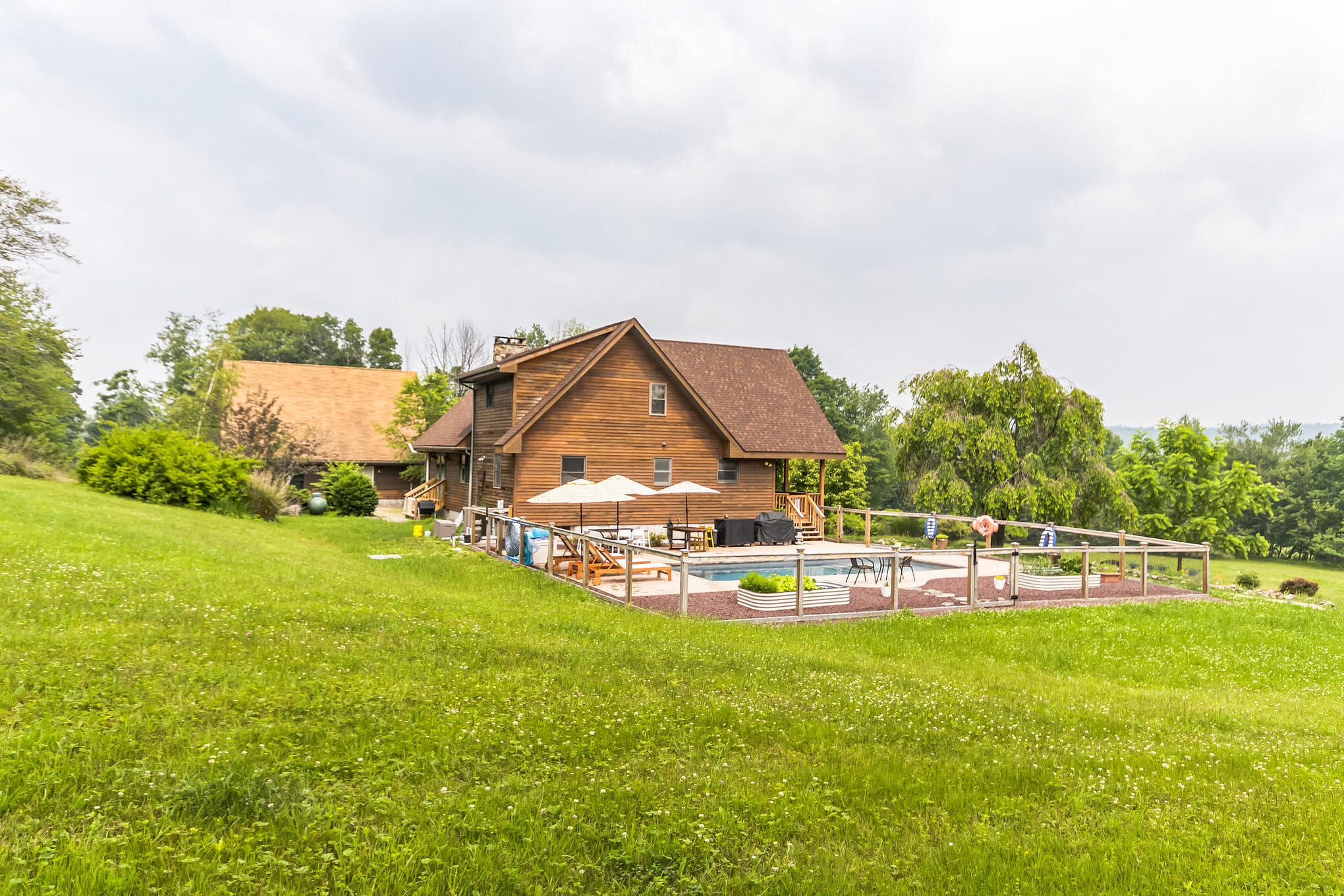 The wooden-sided house is visible from a grassy hillside, with a fenced swimming pool area featuring lounge chairs and umbrellas in the foreground. The expansive green lawn stretches towards distant trees under a cloudy sky.