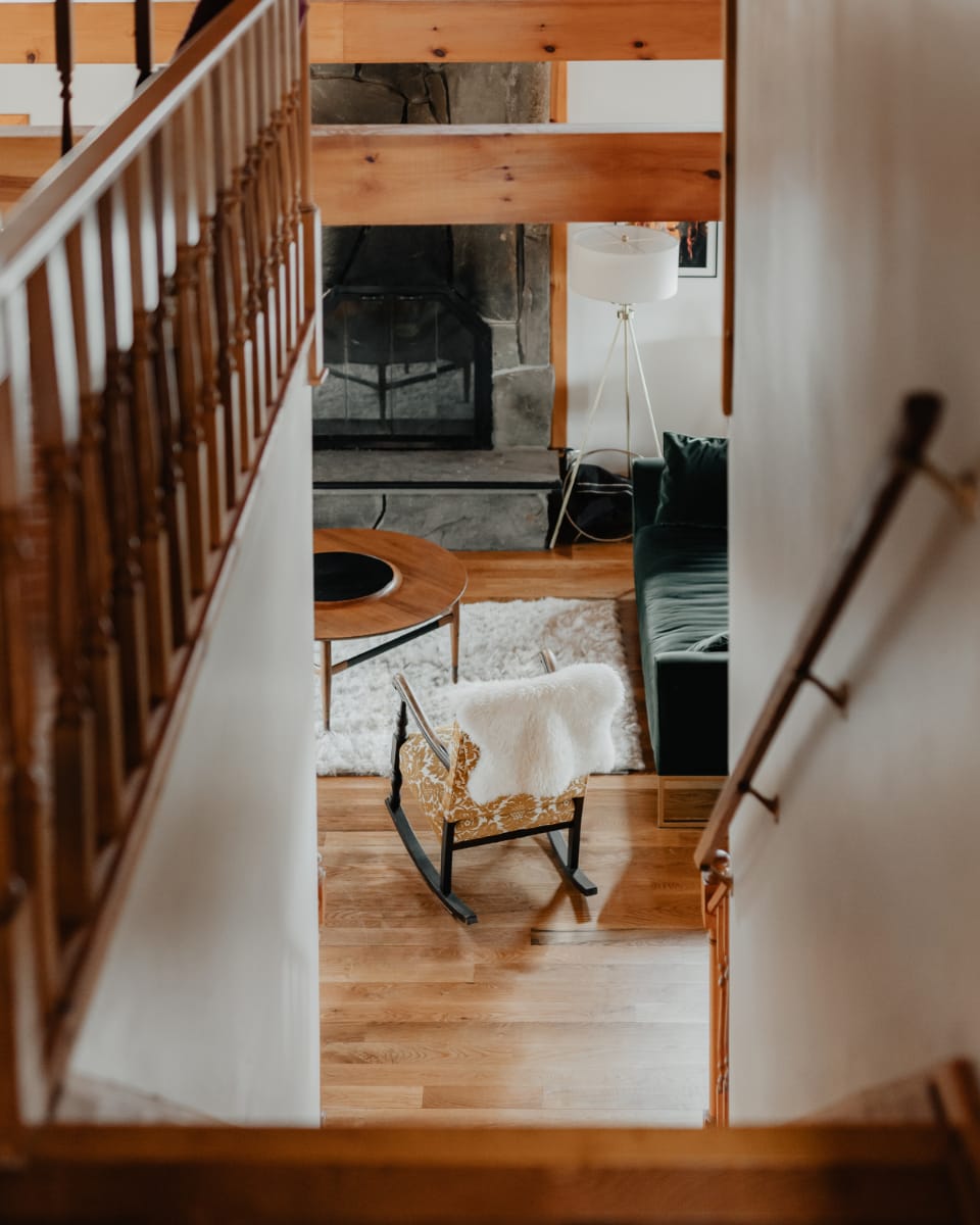 A view down a wooden staircase into a cozy living room with a stone fireplace, a dark green sofa, a round wooden coffee table, and a white shag rug, illuminated by a floor lamp.