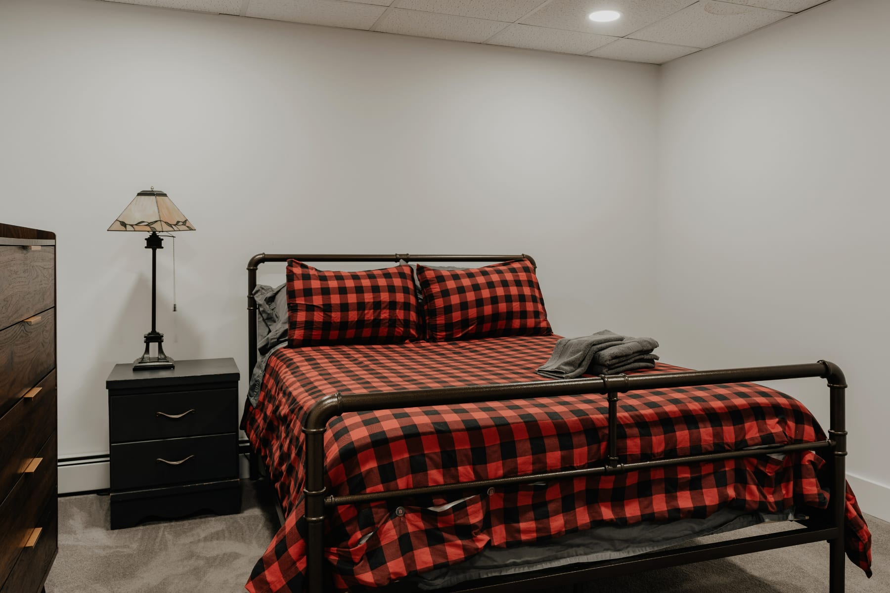 A cozy bedroom with a black metal frame queen bed featuring red and black buffalo check bedding. A black nightstand with a lamp is beside the bed, and the room has light gray carpet and white walls.