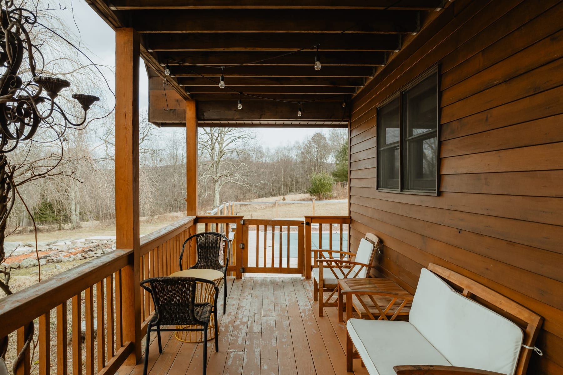 A rustic wooden porch features a dark metal chandelier, string lights, and various outdoor seating arrangements, including wicker chairs and wooden benches with white cushions, overlooking a natural landscape.