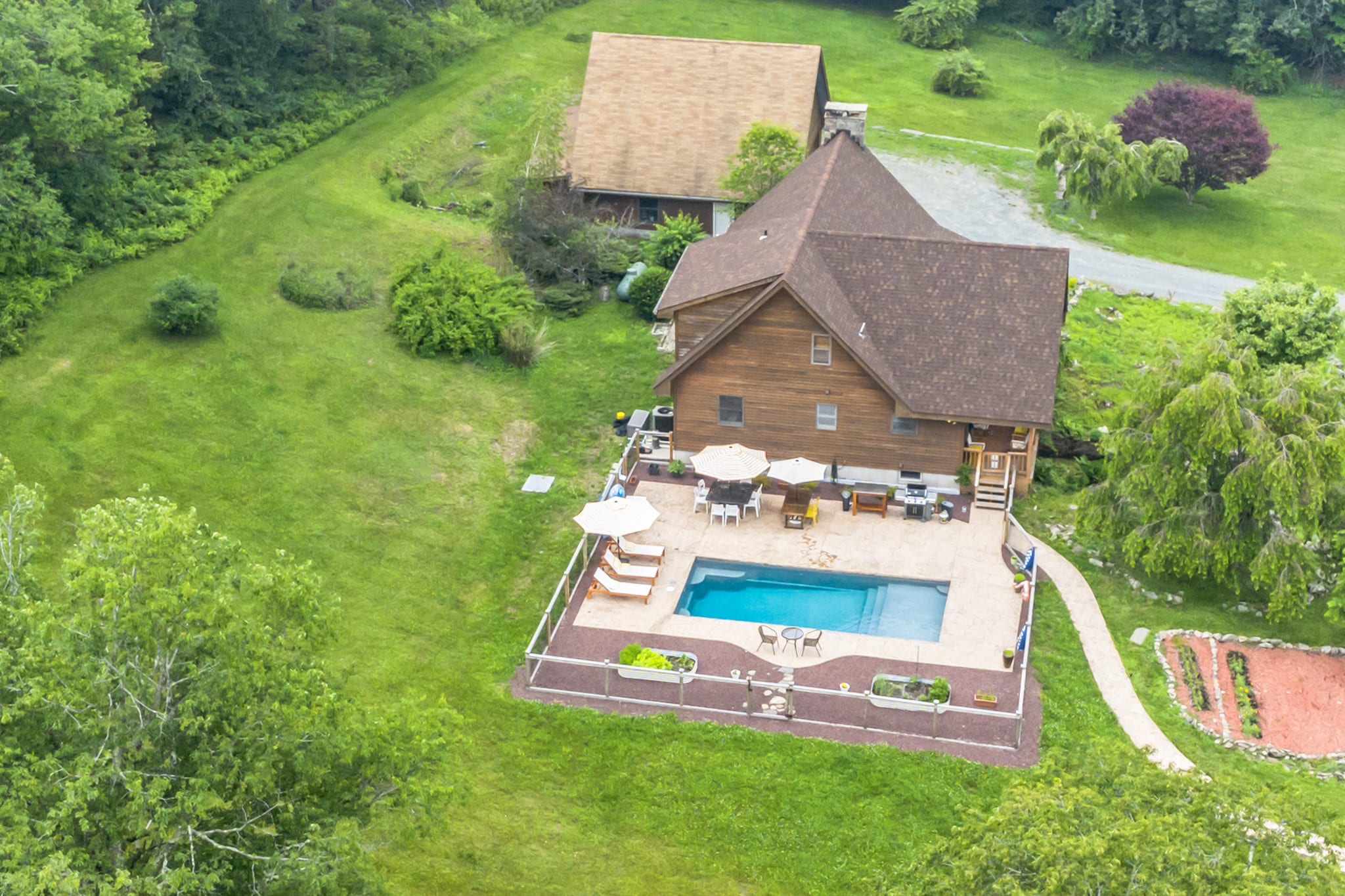 An aerial perspective highlights the back of the wooden-sided house with its brown roof, adjacent to a rectangular swimming pool enclosed by a fence. The pool area is furnished with lounge chairs and umbrellas, set within a lush green landscape.