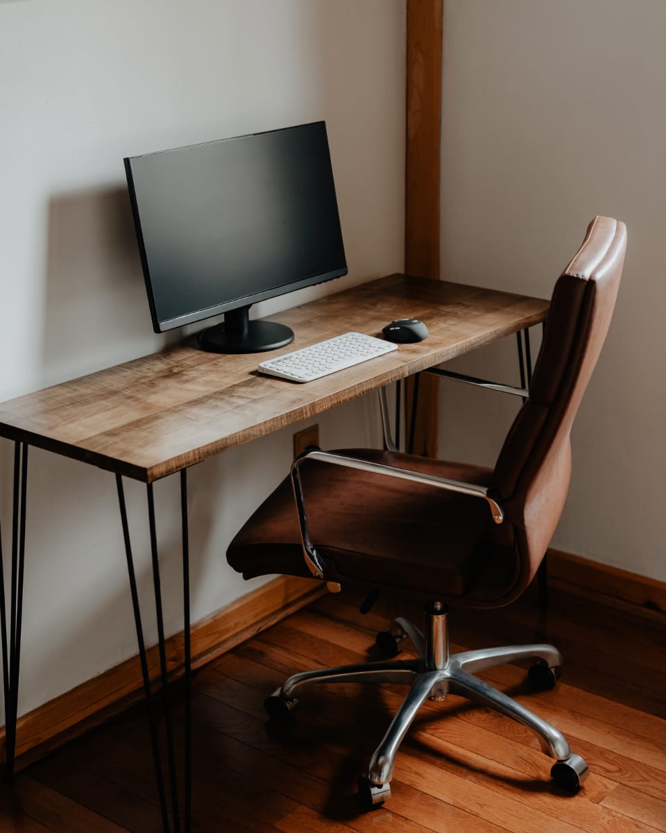 A dedicated workspace featuring a rustic wooden desk with black hairpin legs, a black computer monitor, a white keyboard, and a mouse. A brown leather office chair with chrome accents is positioned in front of the desk, all on polished wooden floors.
