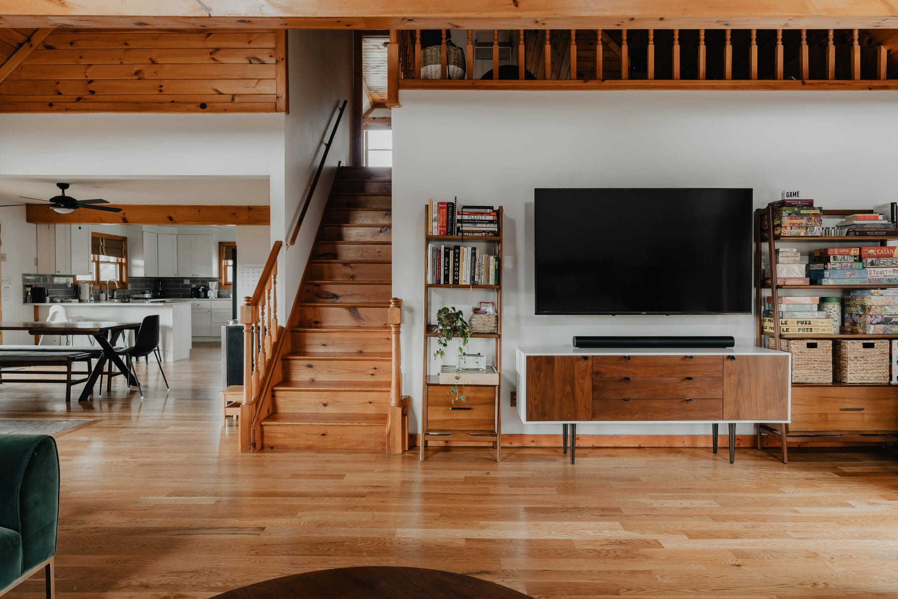 A spacious living area with a large wall-mounted TV above a modern wooden console, flanked by bookshelves filled with books and board games. A wooden staircase leads to an upper level, showcasing the home's open-concept design and warm hardwood floors.
