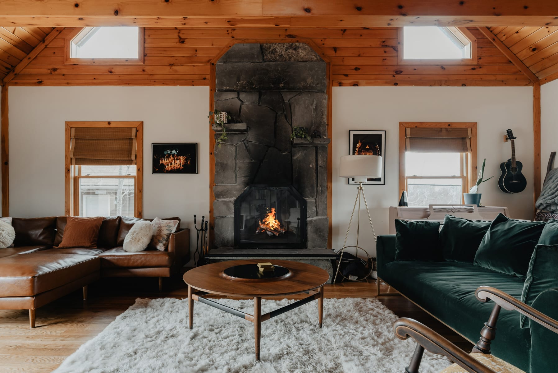 A rustic yet stylish living room centered around a large stone fireplace with a roaring fire. It features a brown leather sectional sofa and a dark green velvet sofa, both facing a round wooden coffee table on a plush white rug.