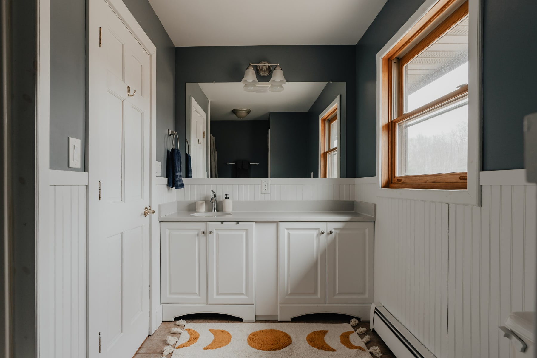 A bright bathroom with dark gray walls and white wainscoting, featuring a white vanity with a large mirror and a window with a wooden frame. A decorative rug with moon phases is on the tiled floor.
