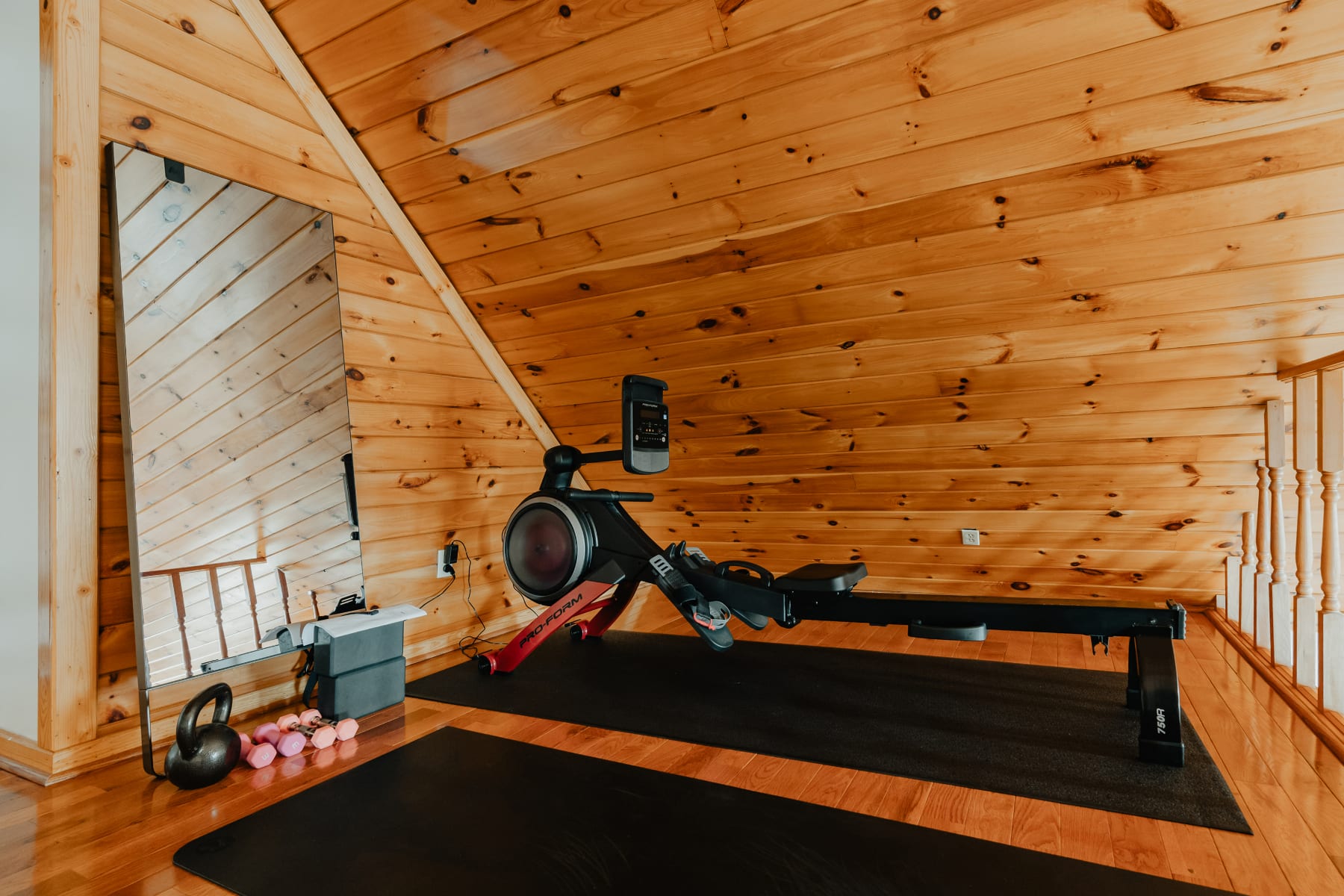Dedicated gym space with a rowing machine, weights, and yoga mats, featuring sloped wood-paneled ceilings and a large mirror, providing an area for fitness activities.