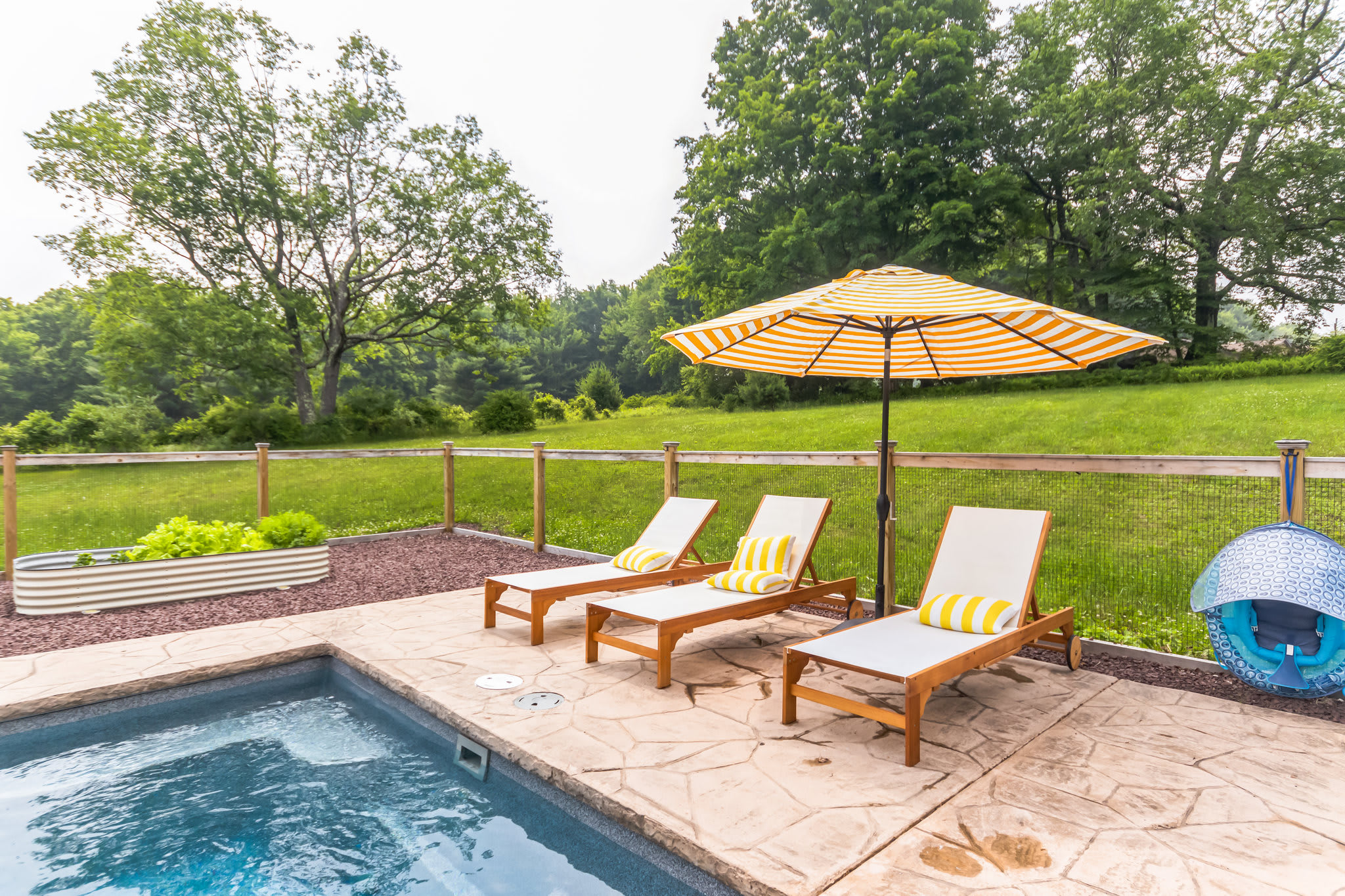 Rectangular inground pool with a stone patio, three lounge chairs with yellow striped cushions, and a matching yellow and white striped umbrella, all enclosed by a wooden fence.
