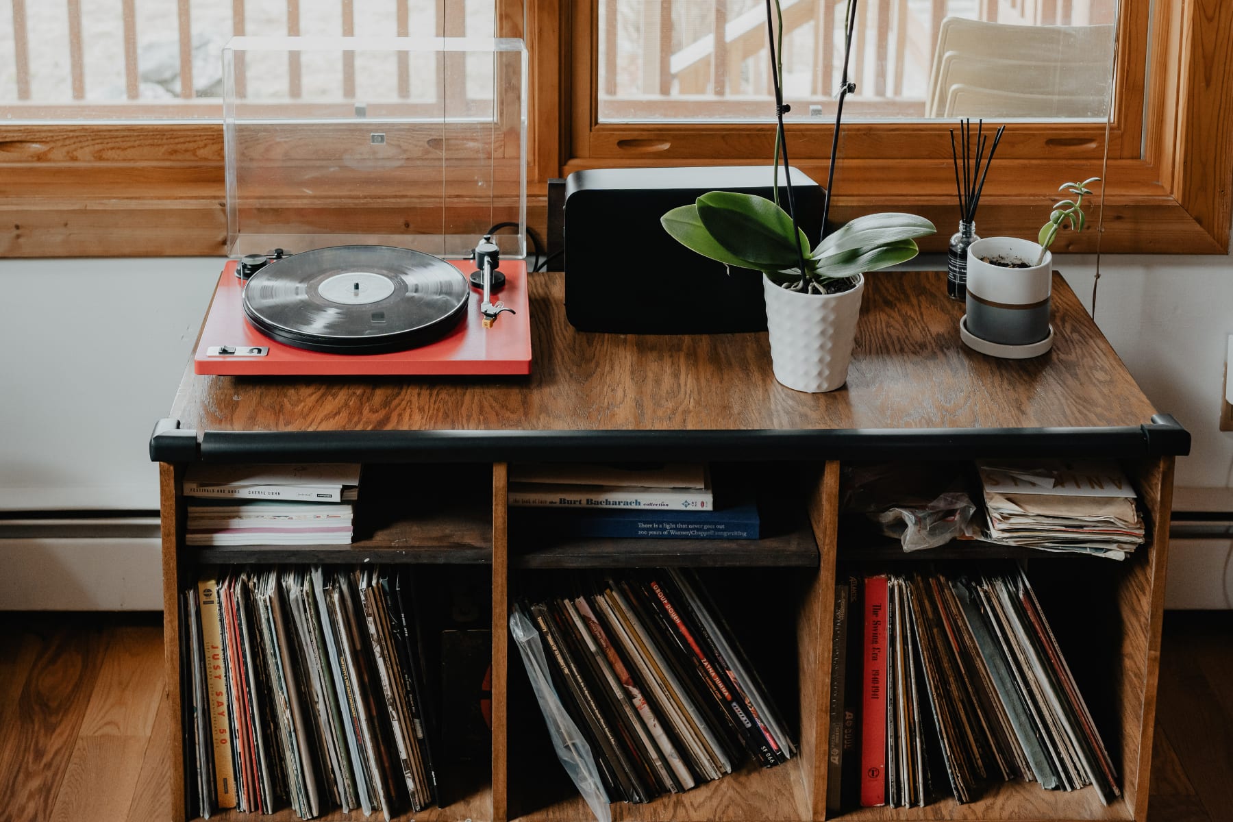 A close-up of a vintage-style record player on a wooden console, surrounded by a collection of vinyl records. A potted plant and a diffuser add decorative touches to this entertainment nook.