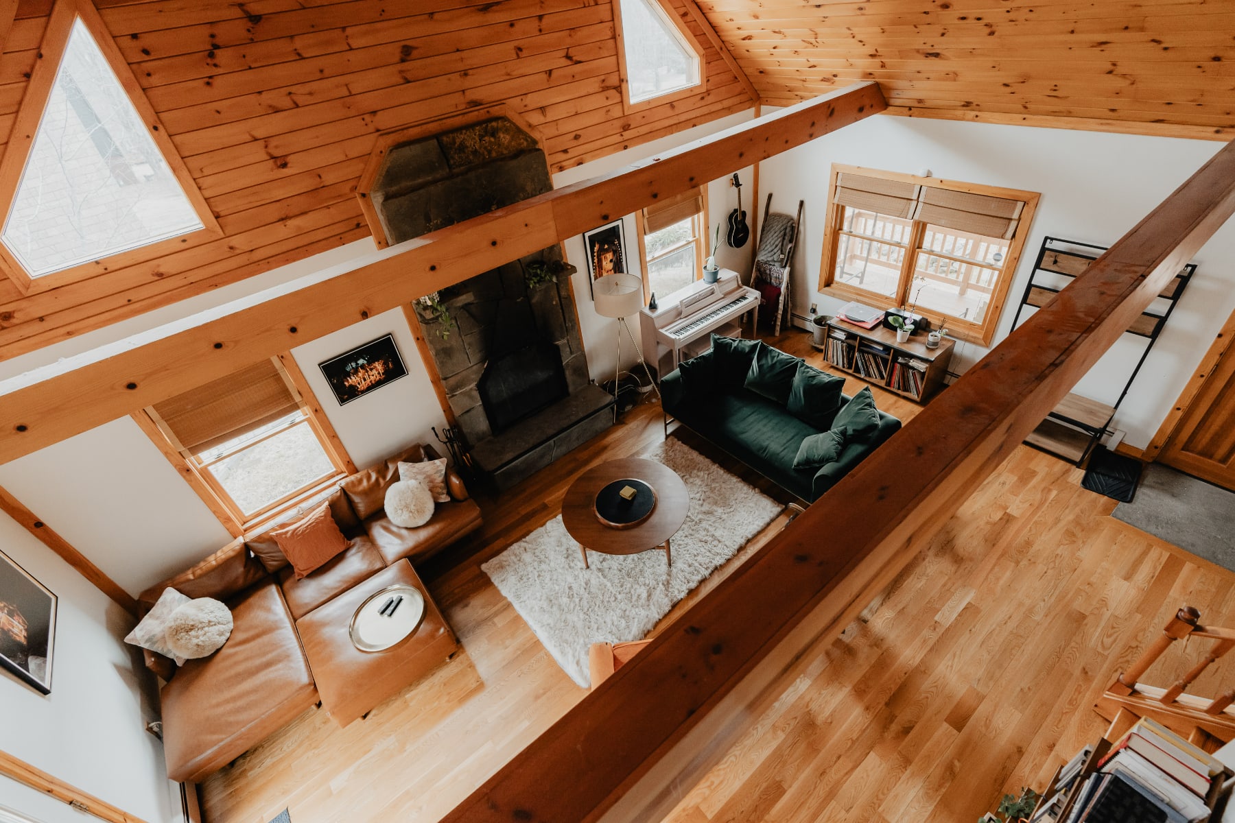 An elevated view of the open-concept living room, highlighting the exposed wooden beams, stone fireplace, and two distinct seating areas with a brown leather sectional and a green velvet sofa. The space is unified by warm hardwood floors and a large white shag rug.