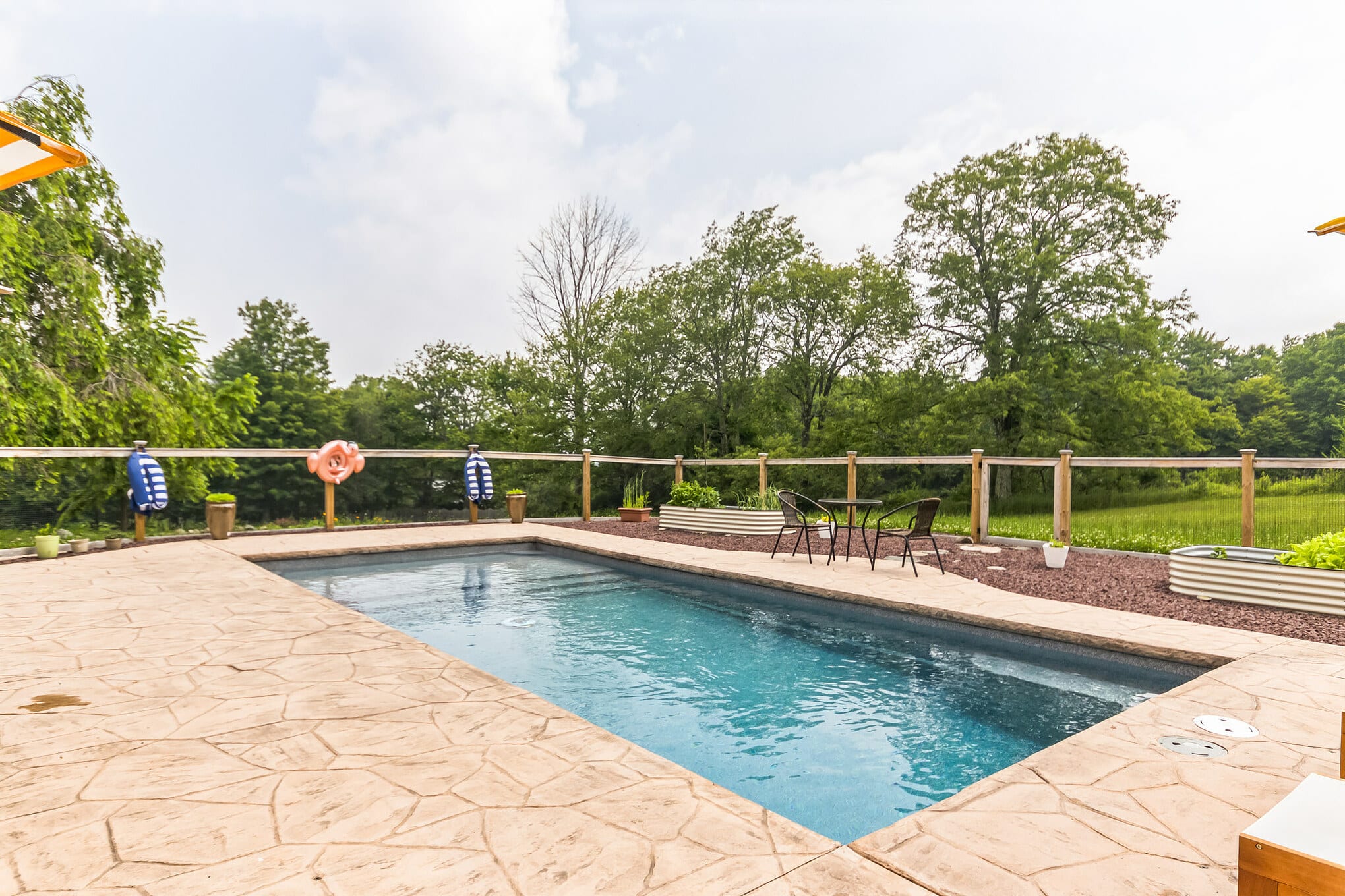 Rectangular inground pool with a textured stone patio, surrounded by a wooden fence and raised garden beds, offering views of a lush green landscape and trees under a cloudy sky.