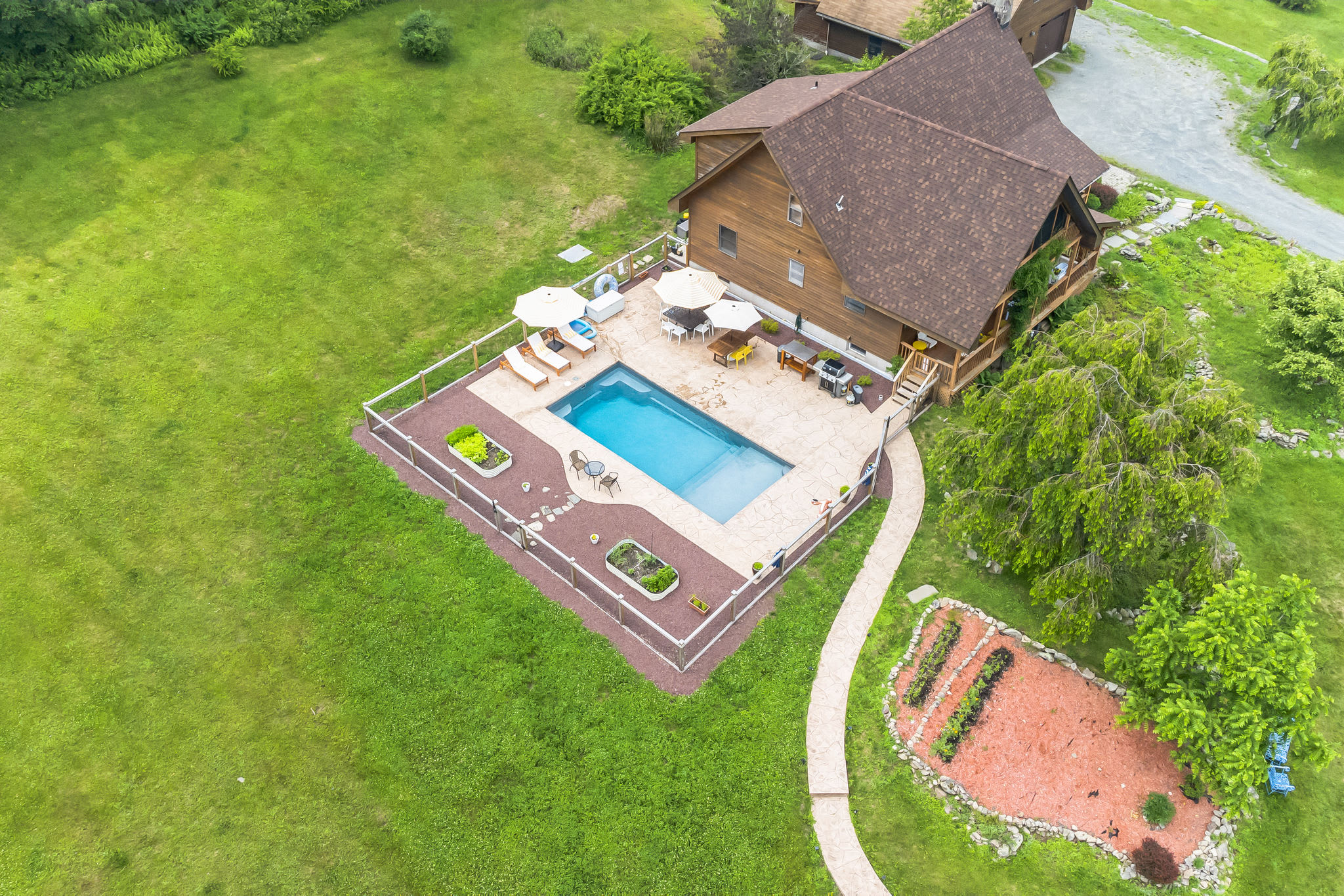 Aerial view of a brown-roofed house with a rectangular inground pool surrounded by a stone patio and fence, set amidst a large green lawn with trees.