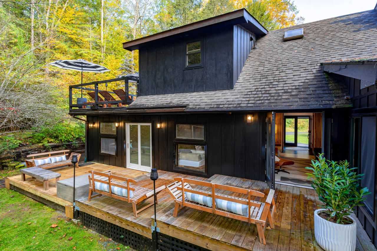 Rear view of the black-sided house with a large wooden deck featuring multiple wooden benches with striped cushions, surrounded by lush green trees and foliage.