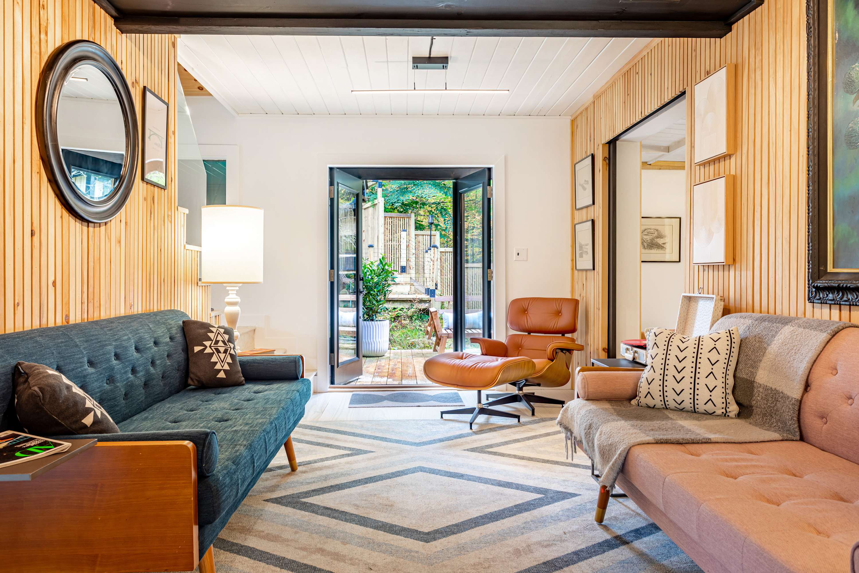 Bright living space with light wood paneling, a blue sofa, a pink sofa, and a brown leather armchair, centered around a geometric patterned rug and leading to an outdoor patio through double doors.