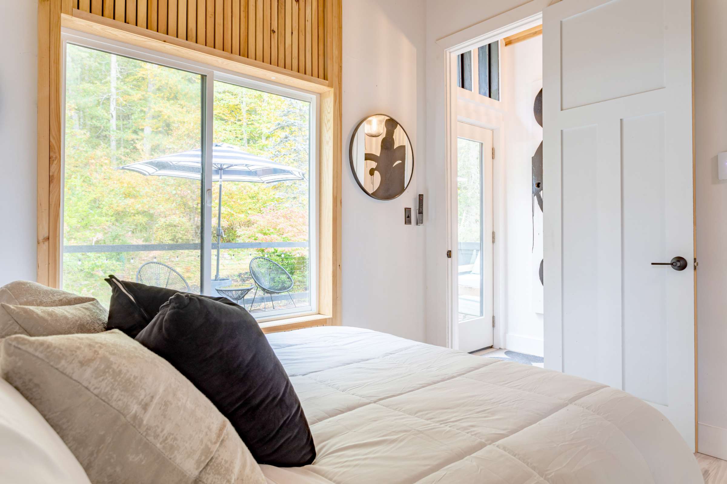 A bright bedroom featuring a bed with dark throw pillows, a large sliding glass door leading to an outdoor area, and a round mirror on the wall. The room has white walls and light wood accents.