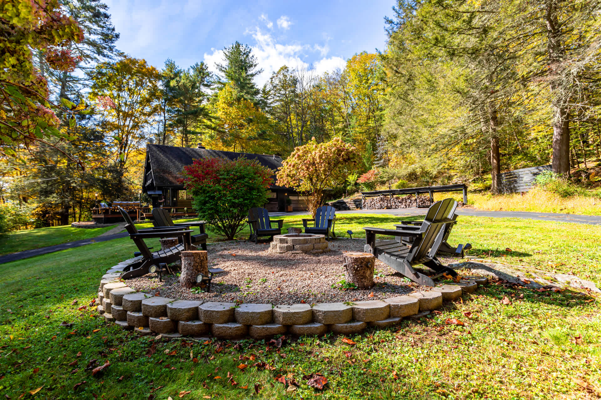 Outdoor fire pit area in a grassy yard, surrounded by a stone retaining wall and several Adirondack chairs. The setting is nestled among trees with autumn foliage, creating a cozy gathering space.
